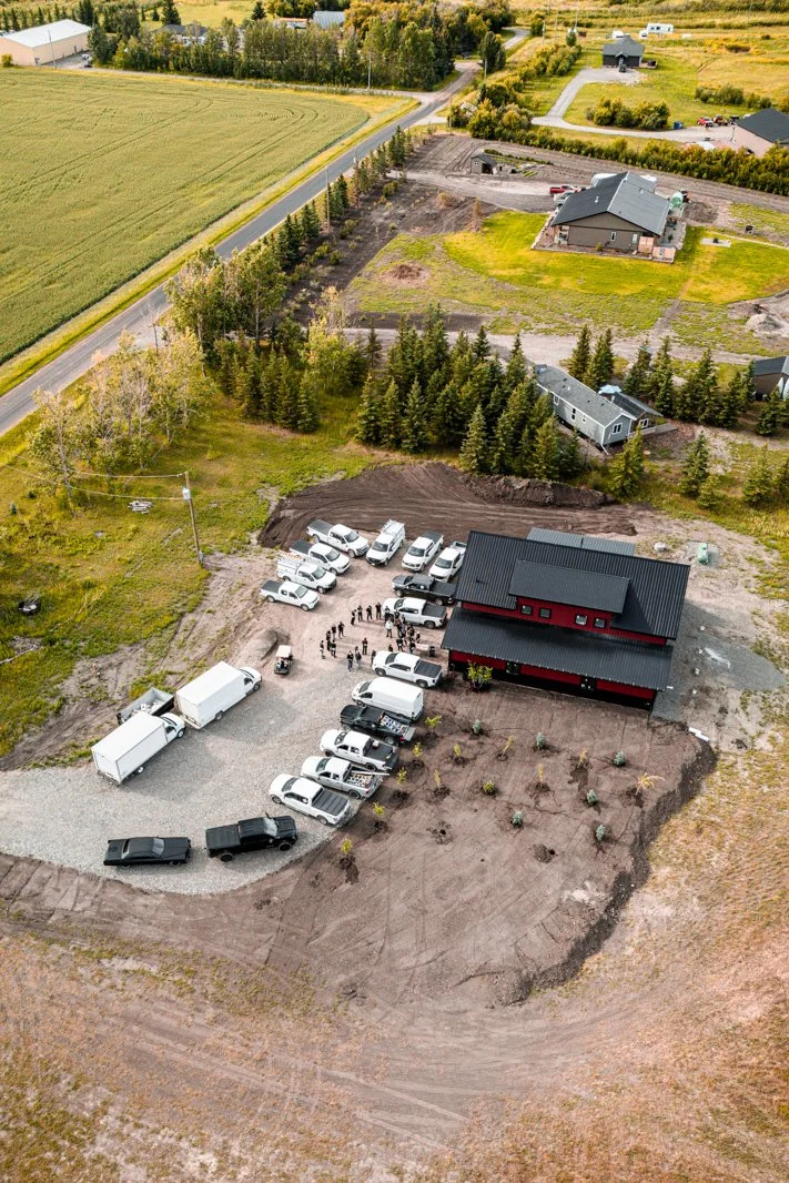 An aerial view of a construction site with a newly built two-story black and red building, surrounded by cars and trucks, with workers gathered outside. The area is partly landscaped with small plants and trees, and there are neighboring houses, tree