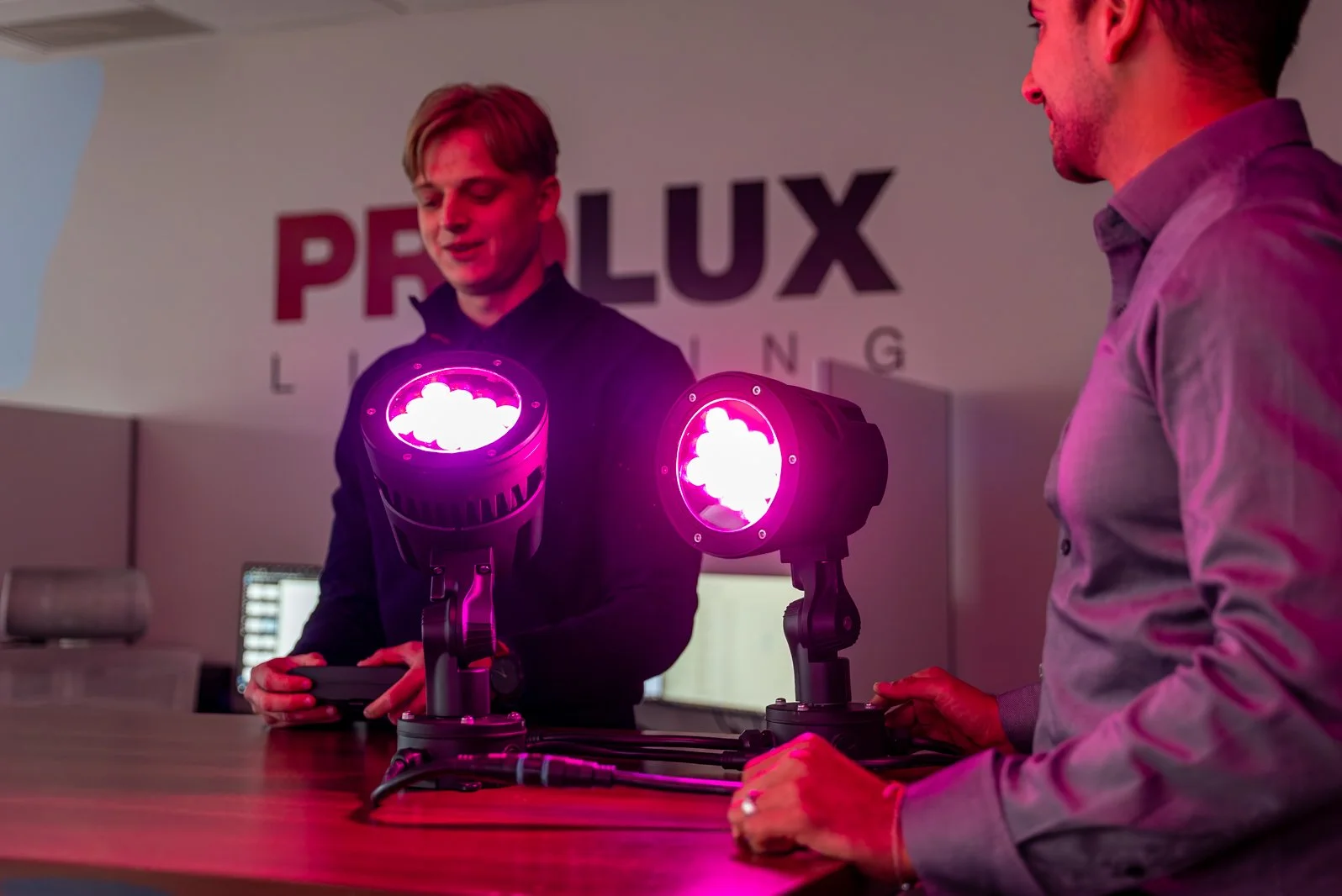 Two men demonstrating lighting equipment with pink lighting at a PRLUX Lighting event.