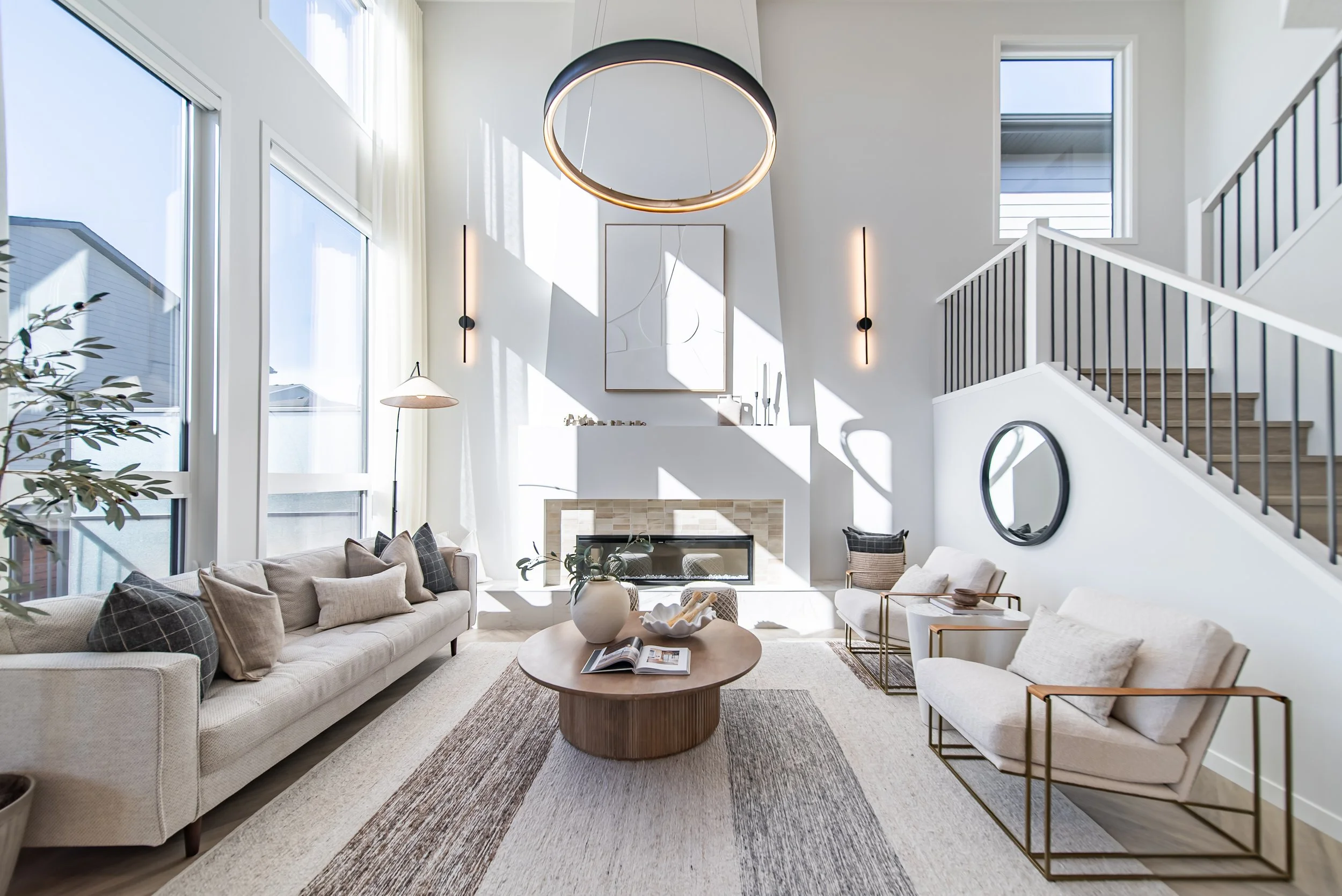 Bright, modern living room with high ceiling, large windows, white walls, and stylish furniture including a beige sofa, white armchairs, and a wooden coffee table. Sunlight streams through windows, creating shadows on the wall.