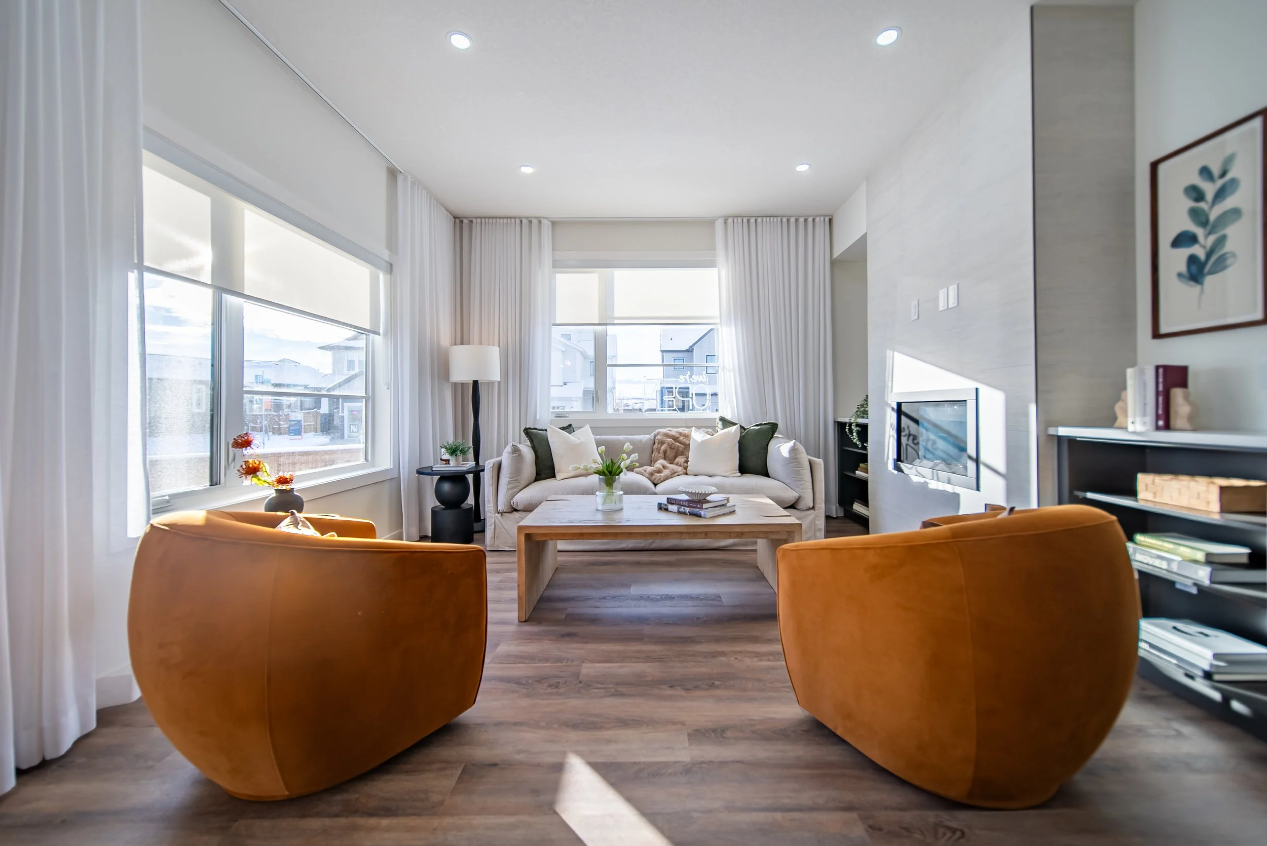 Living room with white sofa, two orange armchairs, wooden coffee table, large windows with white curtains, bookshelf, television, and decorative plants.