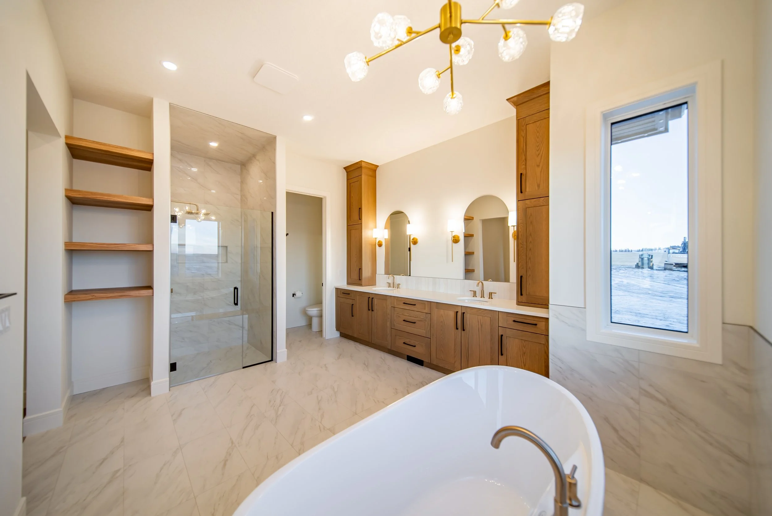 Modern bathroom with wood cabinetry, double sinks, large window showing a waterfront view, separate shower with glass door, bathtub, and bright lighting fixtures.