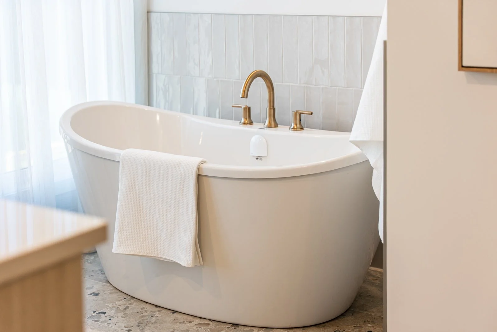 White freestanding bathtub with a cream-colored towel hanging on the side, brass faucet, and white and beige decor in a bright bathroom.