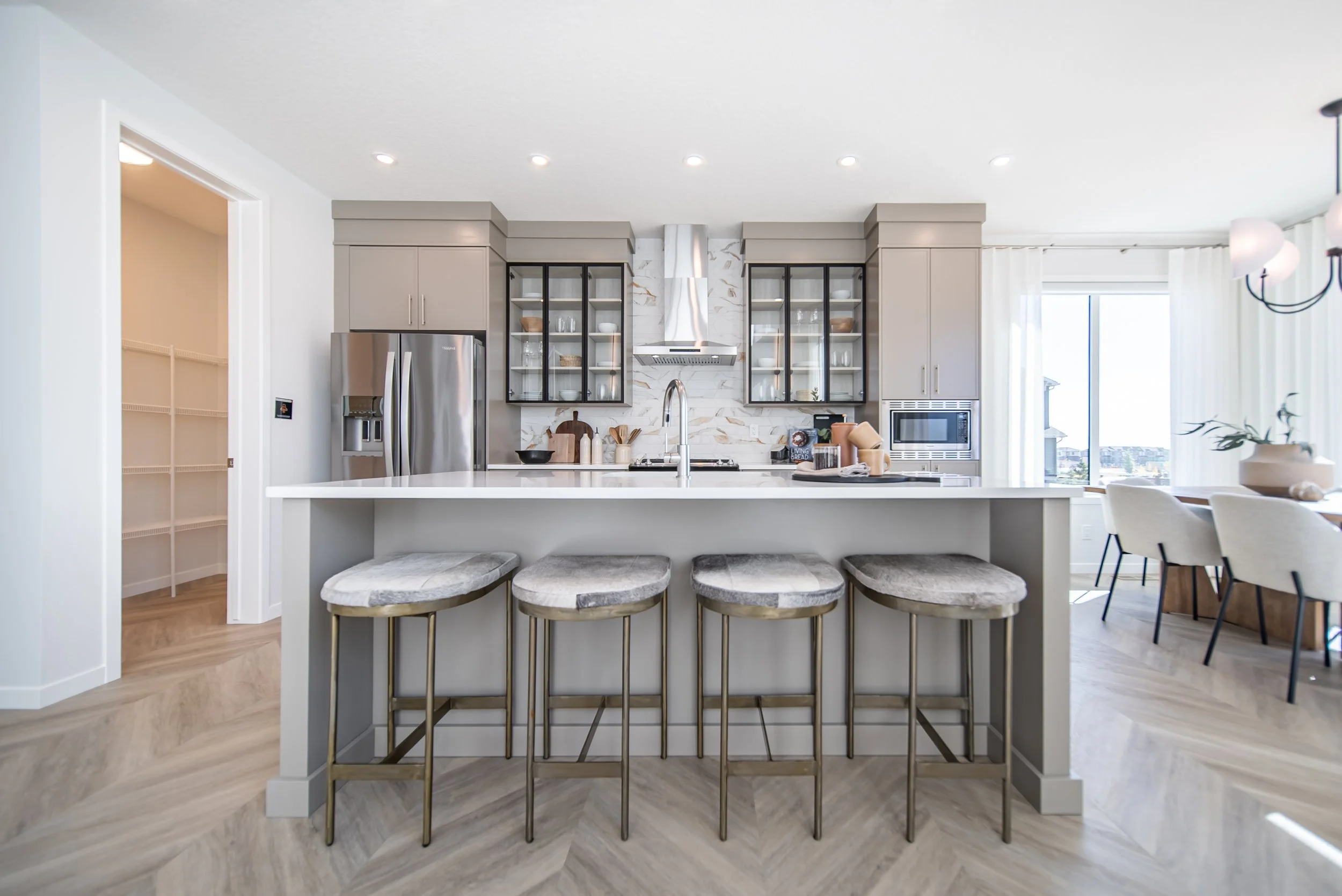 Modern kitchen with island, four upholstered bar stools, stainless steel refrigerator, glass-front cabinets, marble backsplash, built-in microwave, and dining area with large window and white curtains.