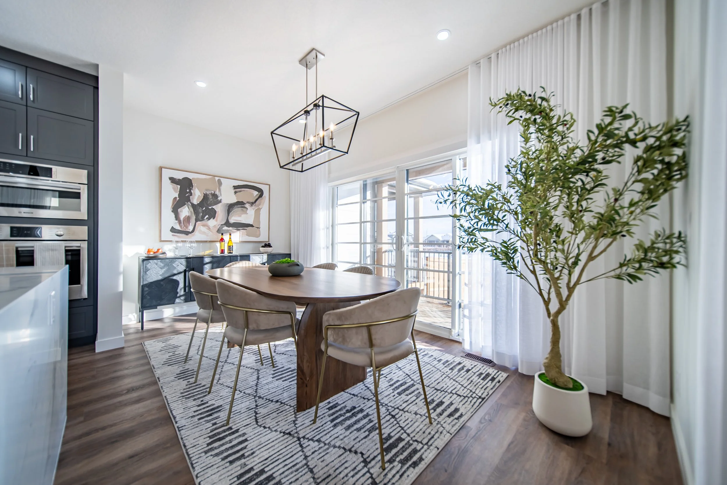 Modern dining room with wooden table, six beige chairs, large potted tree, sliding glass door with white curtains, hanging geometric chandelier, artwork on wall, and sideboard with bottles and bowls.