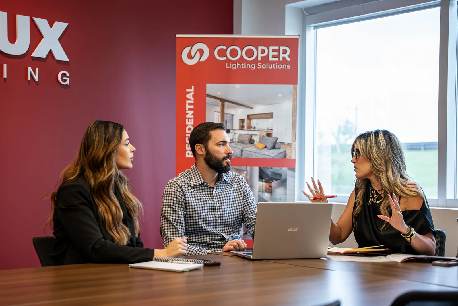 Three people having a business meeting in a conference room with a large window, a laptop, notebooks, and promotional banner in the background.
