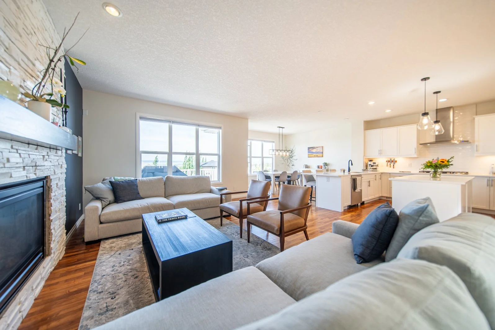 Open-concept living room and kitchen with beige sofas, wooden chairs, a stone fireplace, large windows, hardwood floors, and modern white kitchen cabinets with pendant lighting and a flower vase.