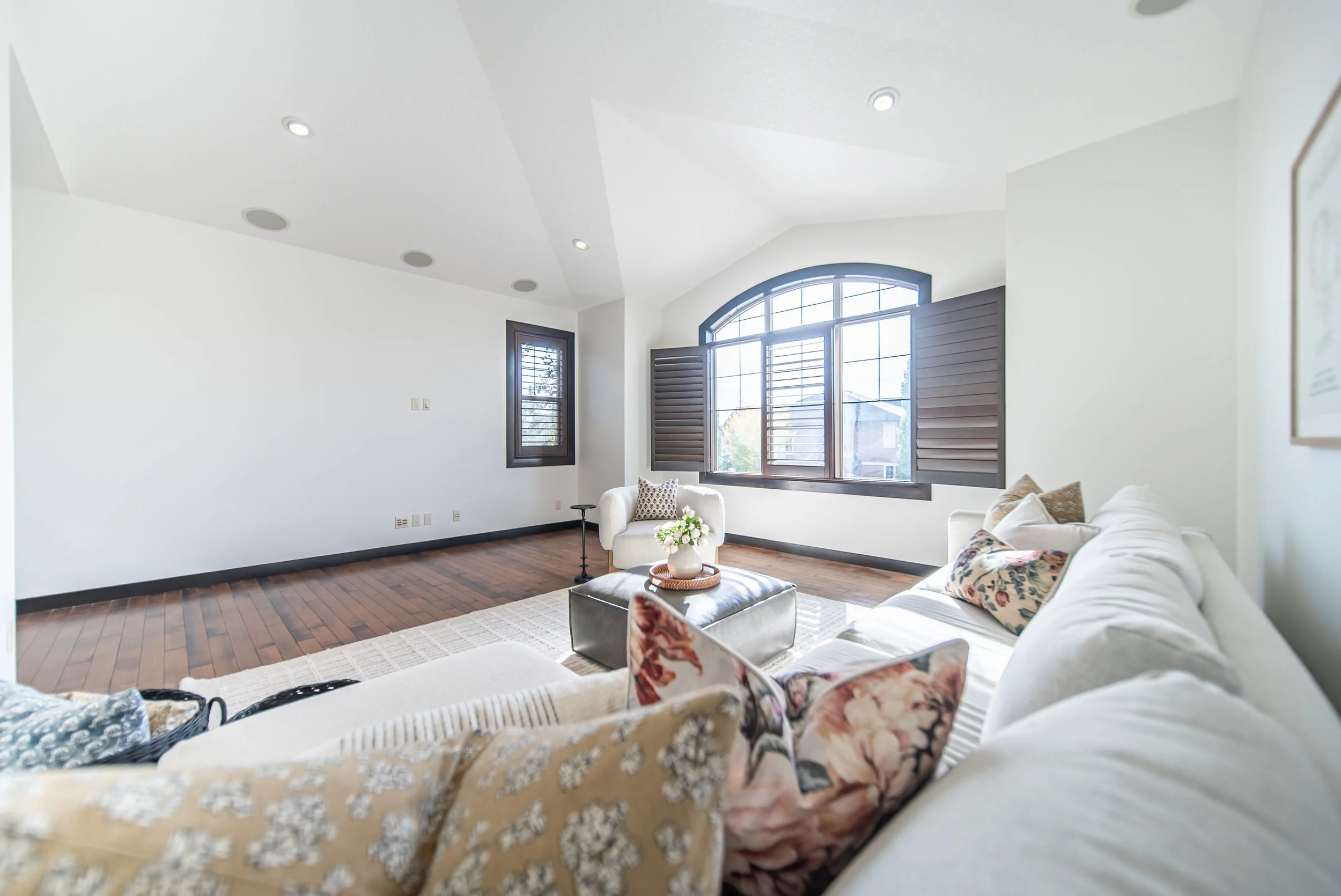 Bright living room with white walls, dark wood flooring, large arched window with shutters, beige sofa with pillows, white armchair, and coffee table with flowers.
