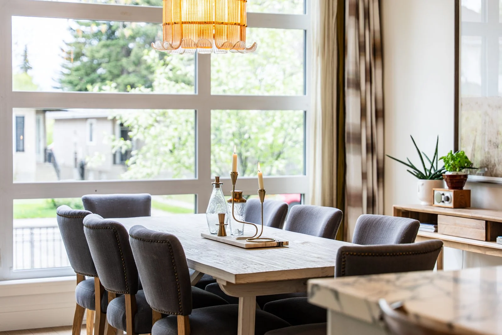 A dining room with a wooden table surrounded by six padded gray chairs. On the table, there are glass bottles and a gold candle holder with three lit candles. Large windows behind the table reveal a view of trees and a neighborhood. To the right, the