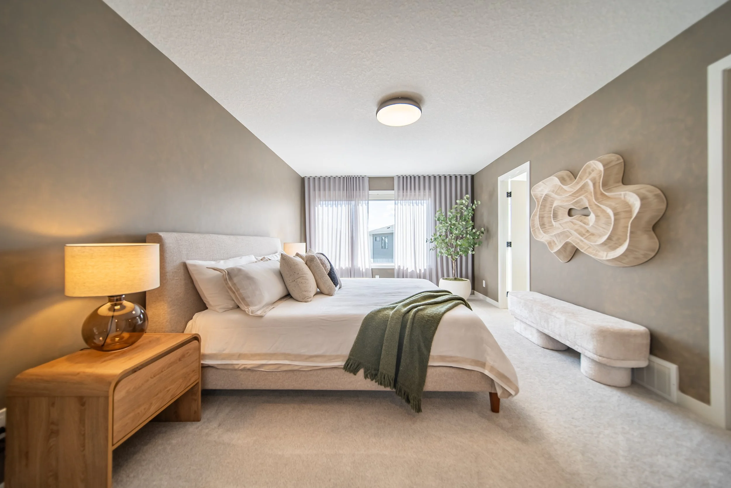 A bedroom with a large bed with pillows and a green throw blanket, a wooden nightstand with a lamp, a large window with sheer curtains, a potted plant, a textured wall decor, and a cushioned bench.