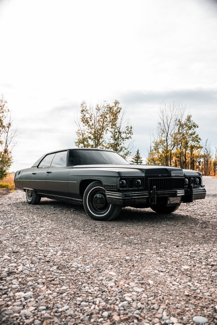 A black vintage Cadillac car parked on a gravel surface with trees and cloudy sky in the background.