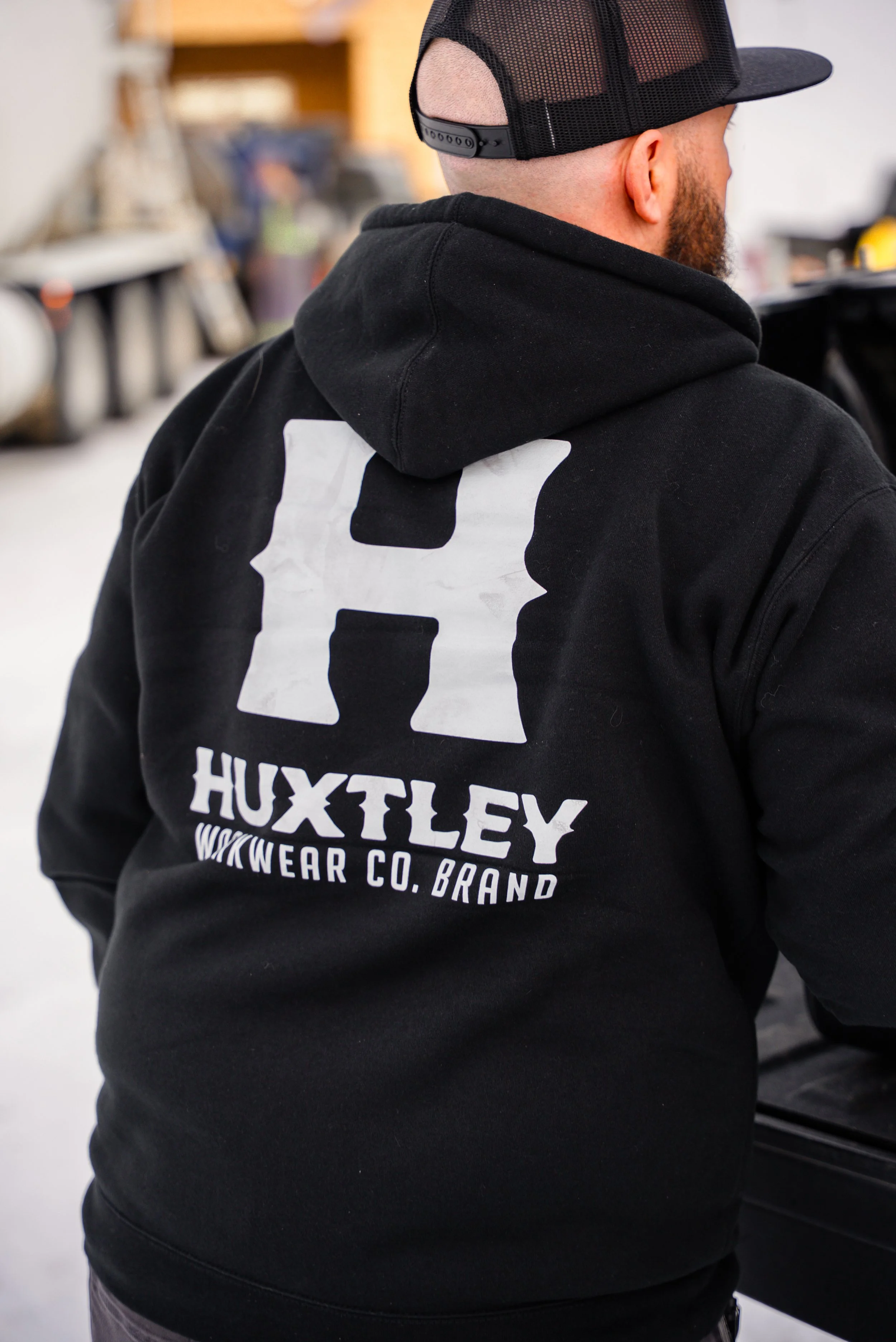 A man wearing a black hoodie with a large white CHP logo, the text 'Huxtley WDWear Co. Brand' on the back, and a black mesh cap, visible from the side in an indoor setting.
