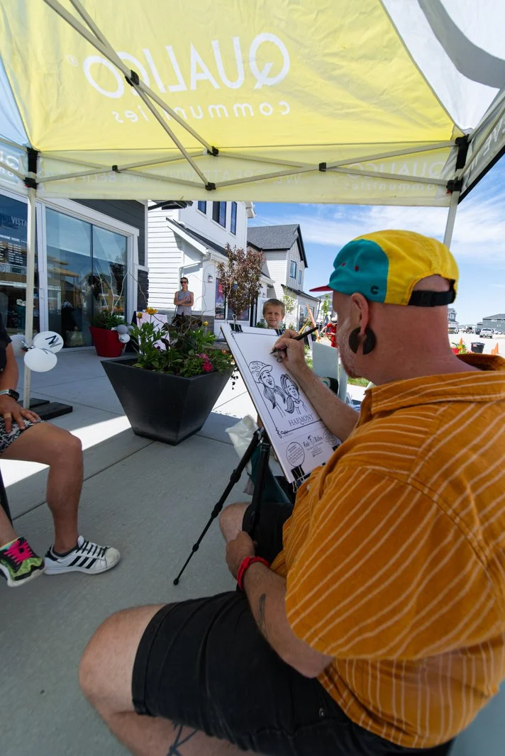An artist in a yellow and blue cap sketches caricatures at an outdoor event under a yellow tent. There are two children sitting for drawings and people standing nearby on a sidewalk with white houses in the background.