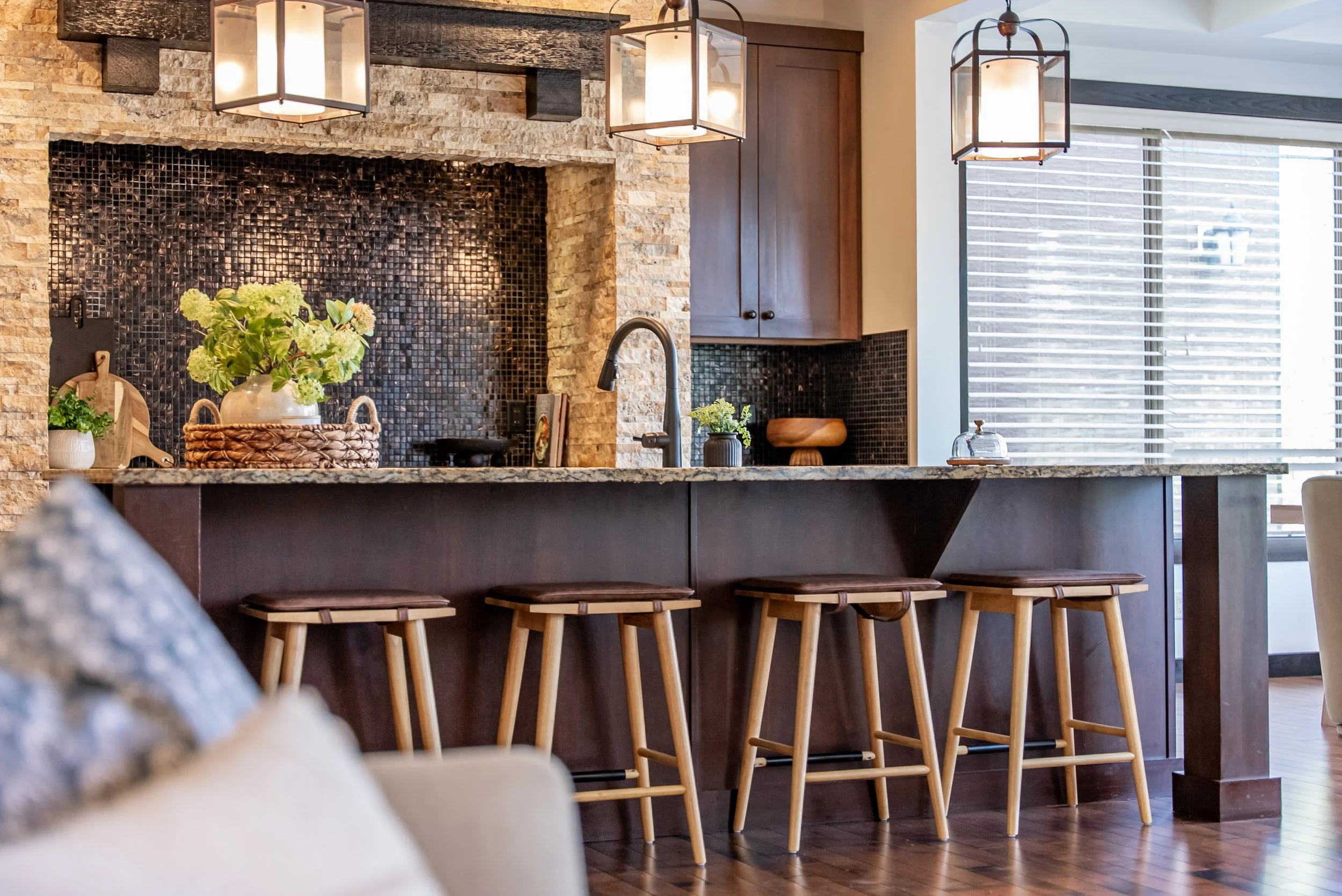 Modern kitchen with dark wooden island, four wooden bar stools, a stone and black tile backsplash, hanging pendant lights, and large windows with blinds.