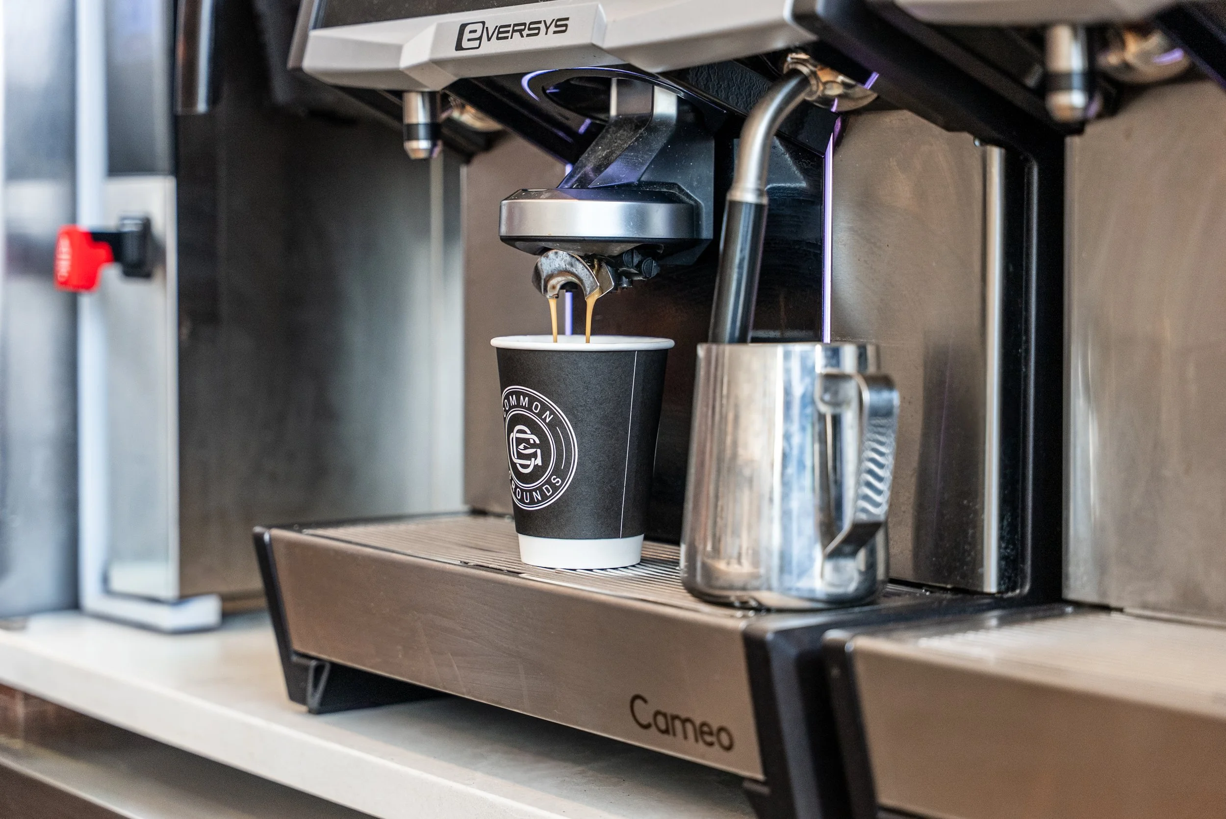A coffee machine dispensing coffee into a black paper cup with a logo, positioned on a metal drip tray, with a stainless steel milk frothing pitcher nearby.