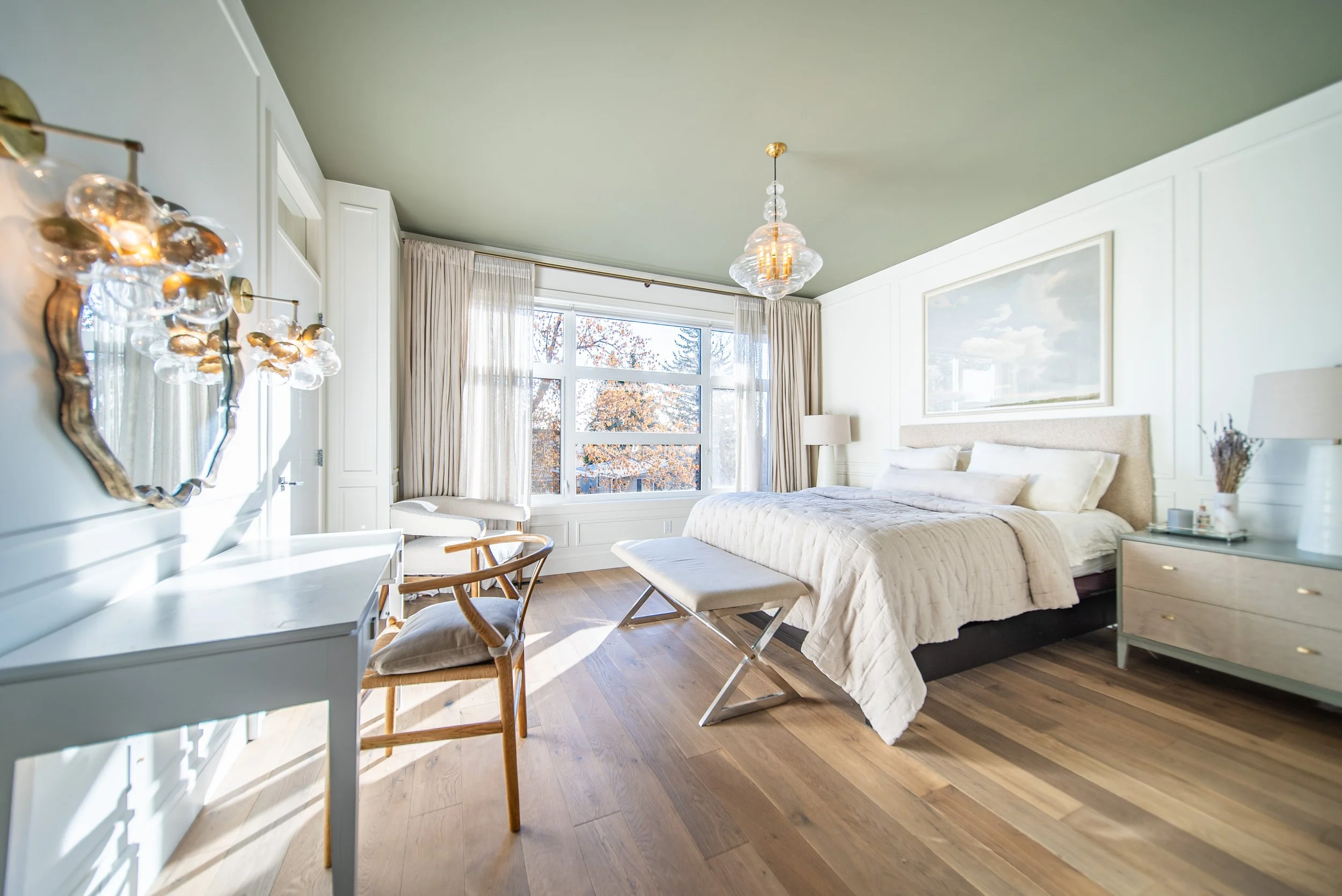 Bright bedroom with large window, green ceiling, white walls, and hardwood floor. Features a bed with beige bedding, a dresser with lamps, a bench at the foot of the bed, and a chair near a white desk with a decorative mirror.
