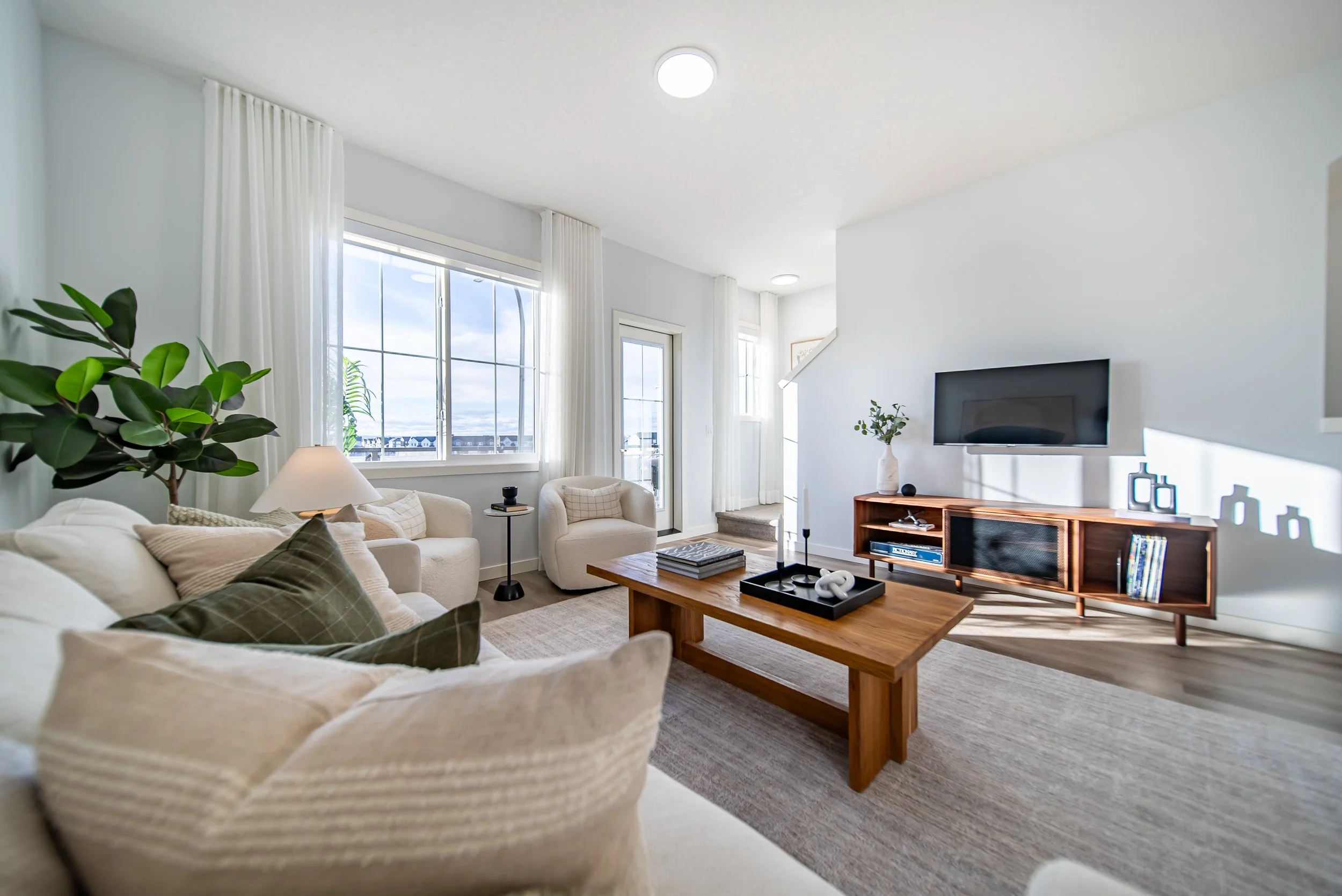 Living room with white sofa, beige armchairs, a wooden coffee table, a flat-screen TV, large windows with white curtains, and decorative items.