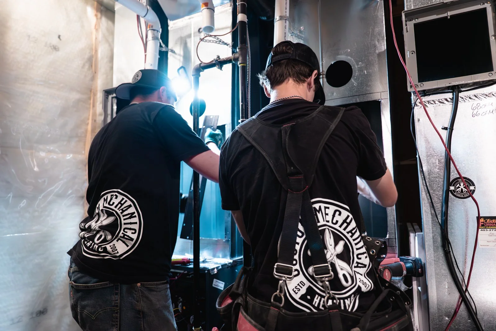 Two individuals working on a mechanical or electrical project inside an industrial setting, wearing black shirts with a logo that says "Mechanical" and "Heinz".