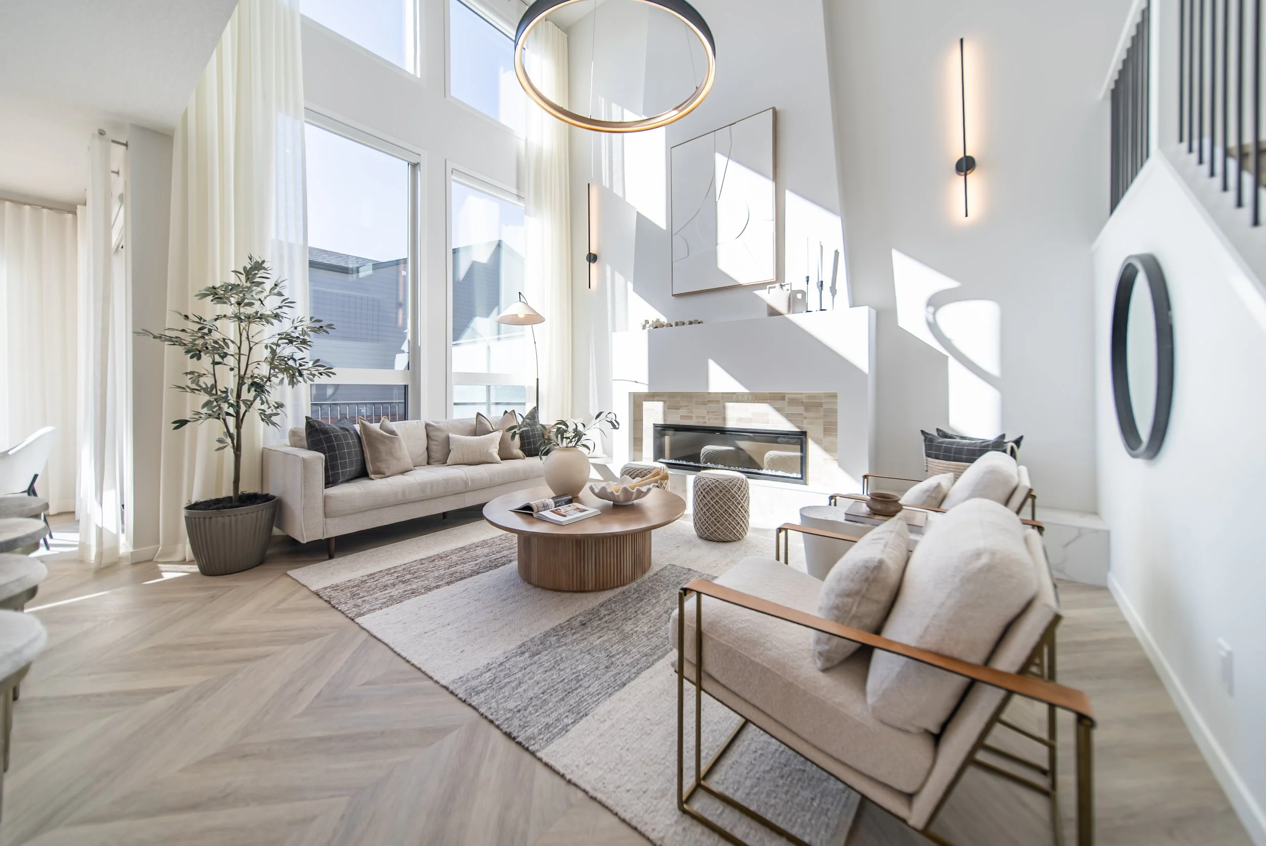 Bright living room with large windows, neutral-colored sofas, a wooden coffee table, modern fireplace, tall indoor plant, and contemporary wall lighting.