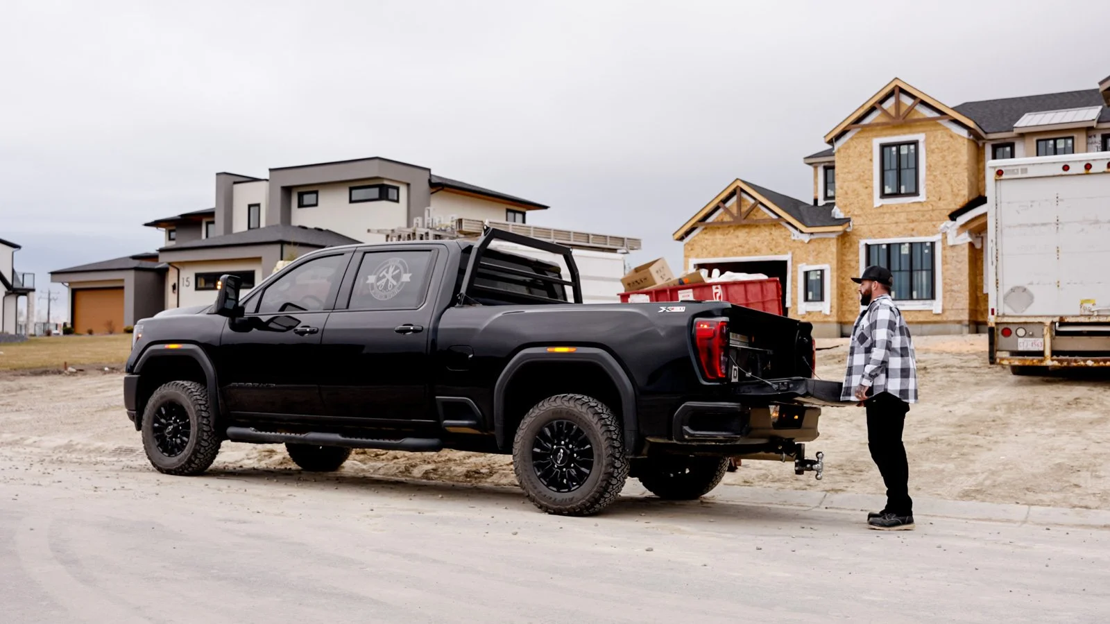 A man in a plaid shirt standing beside a black pickup truck at a construction site with houses in the background.
