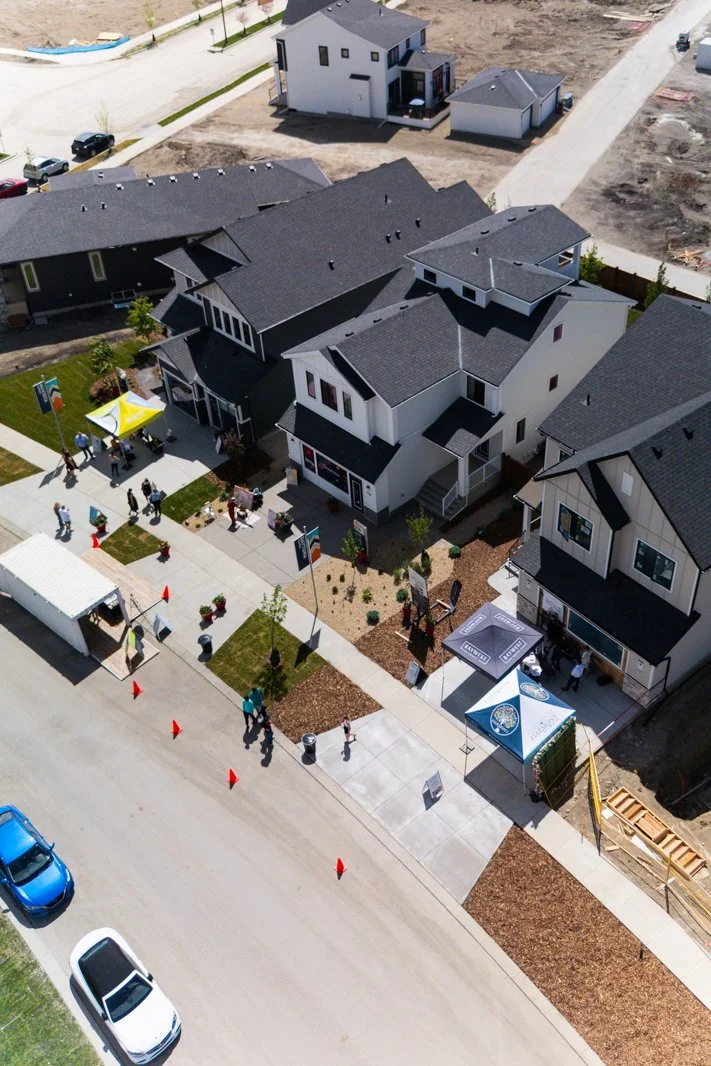 Aerial view of a small community event with people walking on a sidewalk, tents, and small trees in front of modern houses on a residential street.