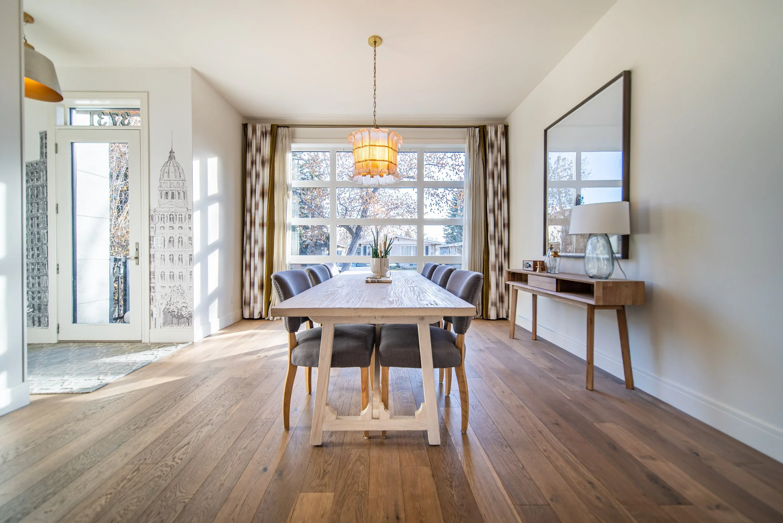 Dining room with large window, wooden table, eight upholstered chairs, and a wooden sideboard with a lamp and decorative items.