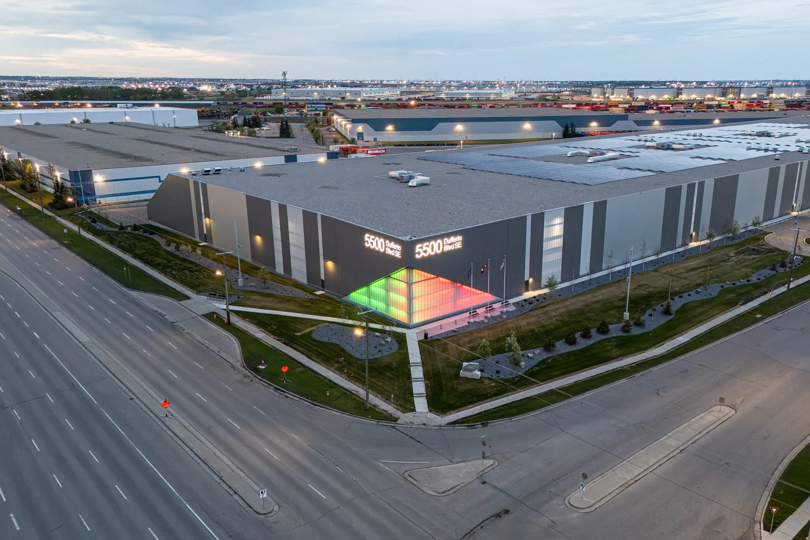 An aerial view of a large industrial building at dusk, with the address 5500 Dufferin Blvd SE displayed on its side. The building has a modern design with a colorful illuminated entrance area, and the surrounding area includes roads, sidewalks, small