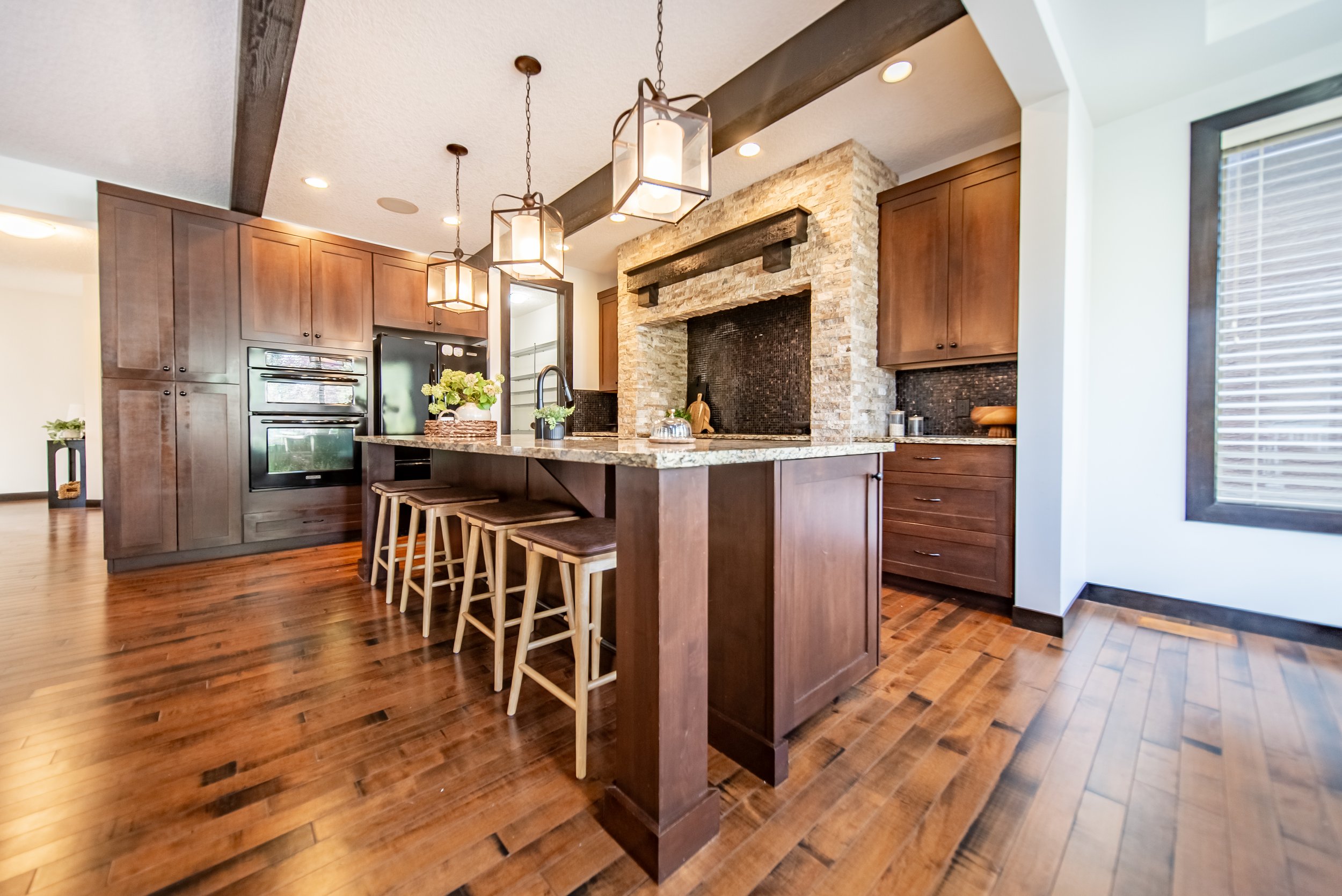 Modern kitchen with dark wood cabinets, a stone fireplace feature, granite countertops, wooden flooring, and three hanging pendant lights above an island with four stools. There is a window with blinds on the right.
