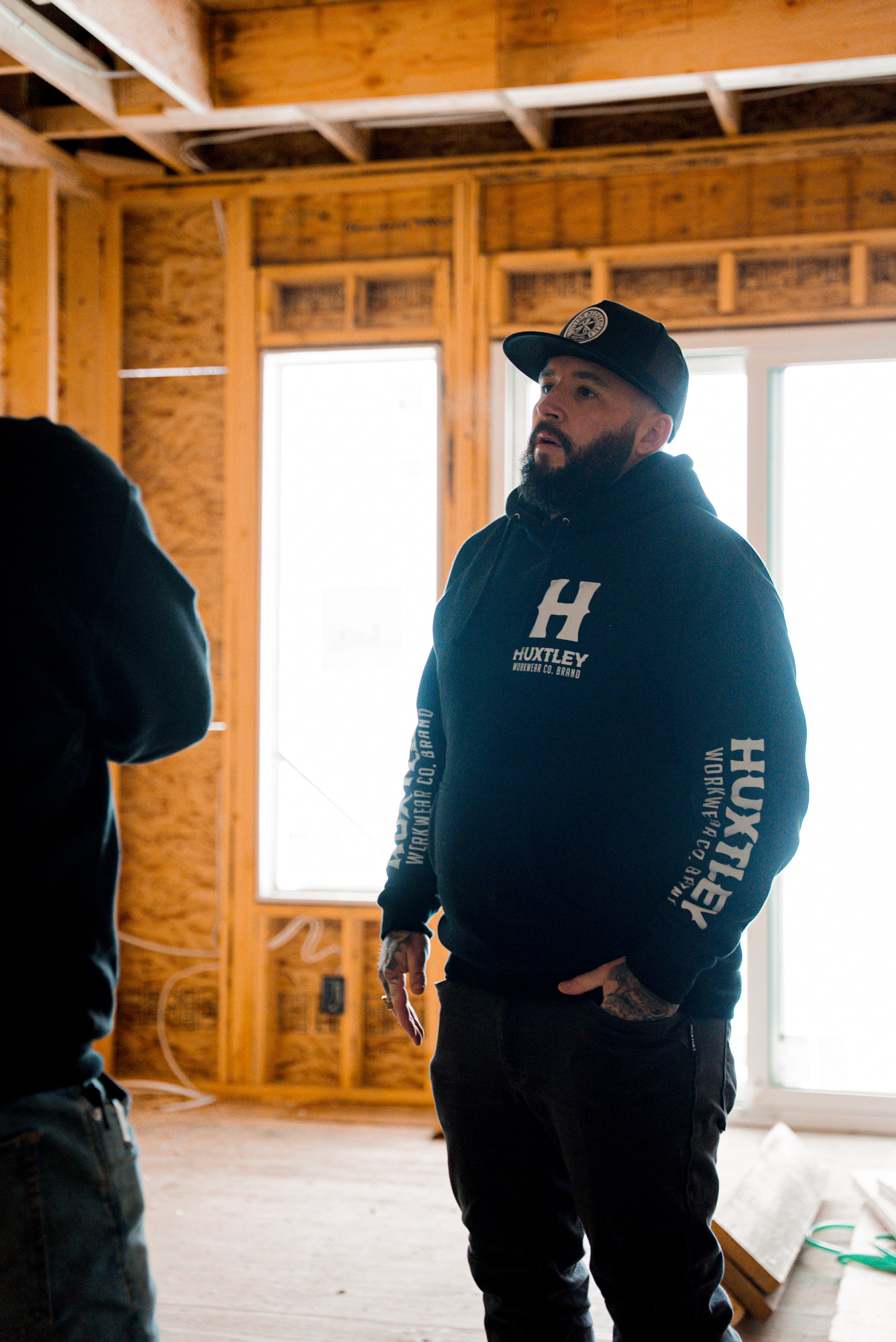 Man with a beard wearing a black hoodie and cap, inside a wooden building under construction.
