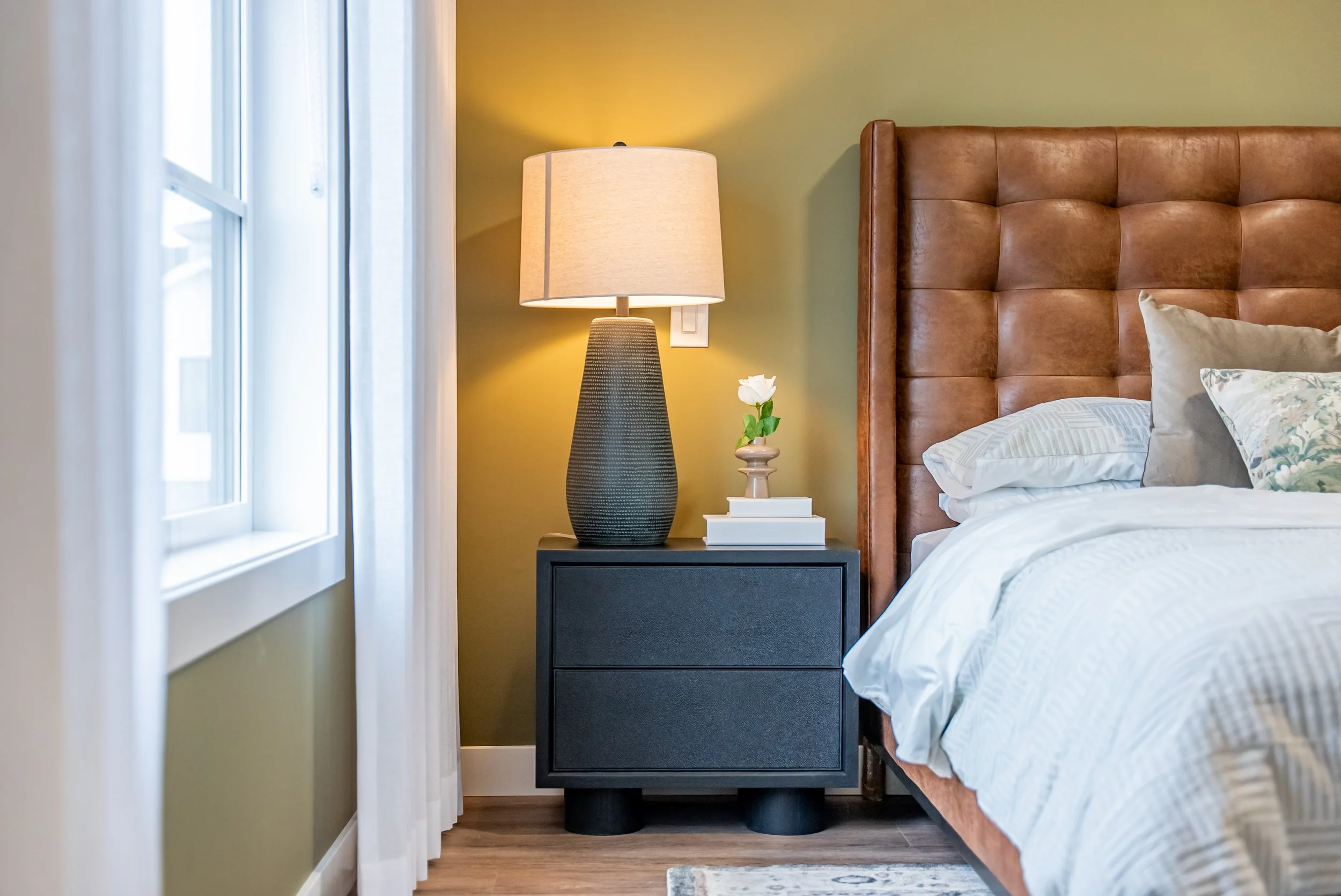 Side view of a bedroom featuring a black nightstand with a tall lamp, a small stack of books, and a white flower in a vase, next to a bed with a brown upholstered headboard, white bedding, and decorative pillows, with a window and white curtains in t