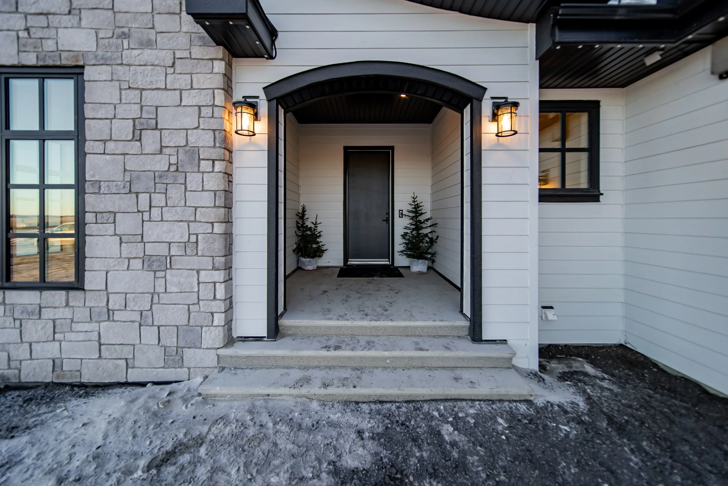 Front entrance of a modern house with stone and white siding, black door, wall-mounted outdoor lights, two small Christmas trees in pots, concrete steps, and a snow-dusted ground.