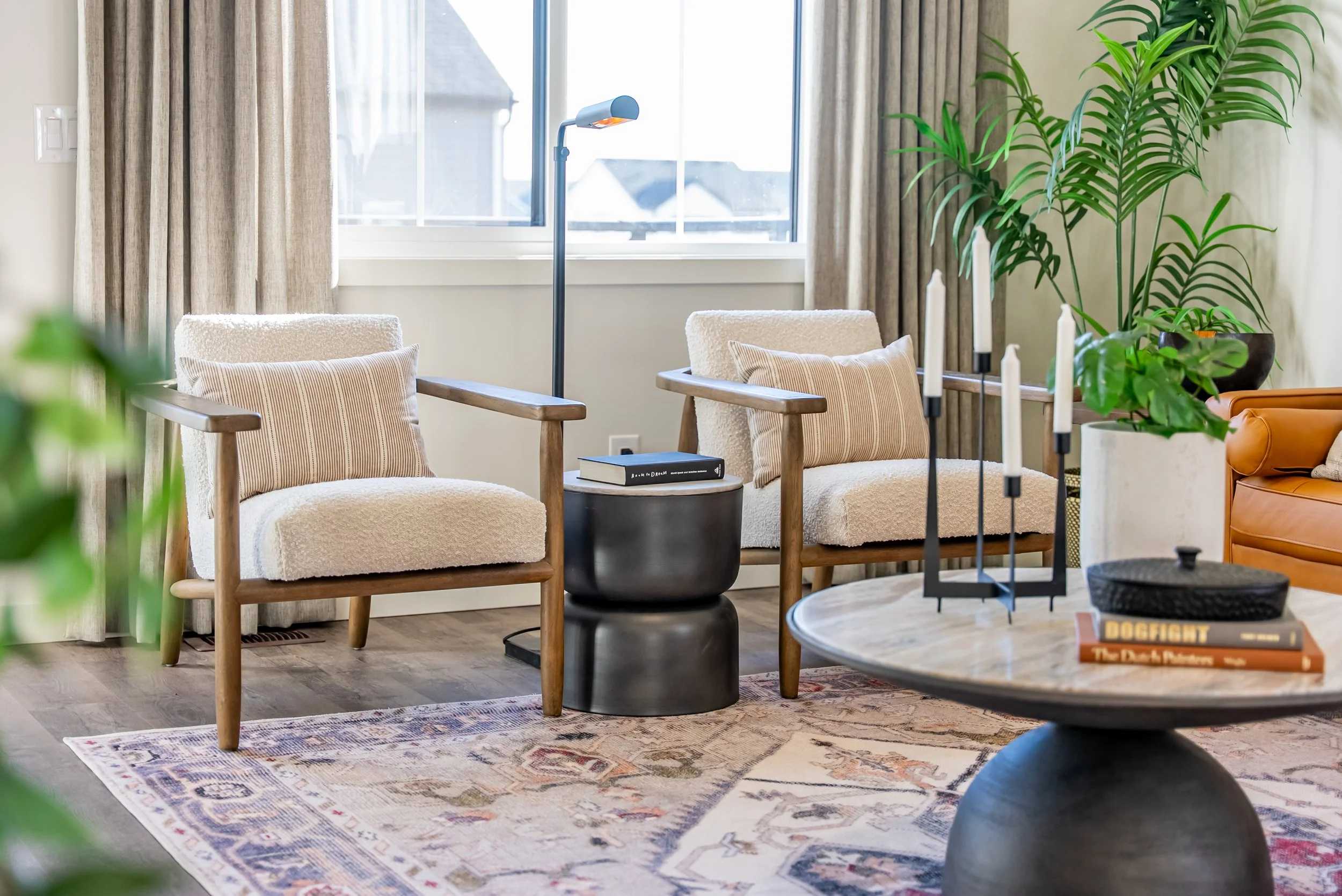 Living room with two beige armchairs, a round wooden coffee table, large green potted plants, books, and candles, near a window with curtains.