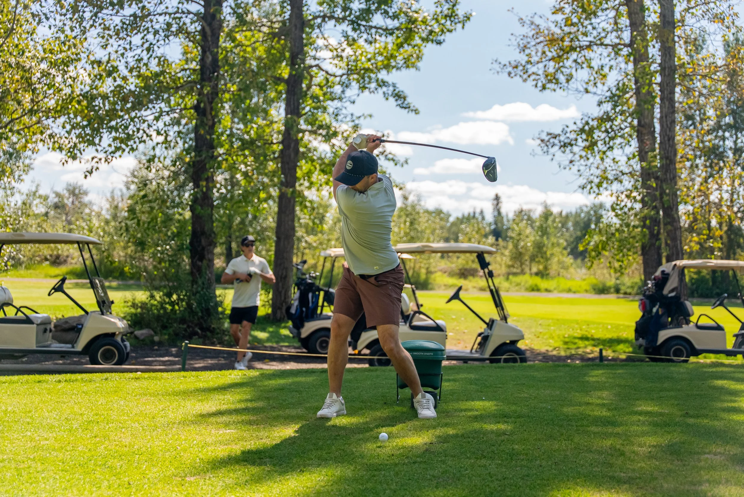 A man playing golf on a golf course, preparing to hit the golf ball with a club, with a golf cart and another person in the background, surrounded by trees and a blue sky.