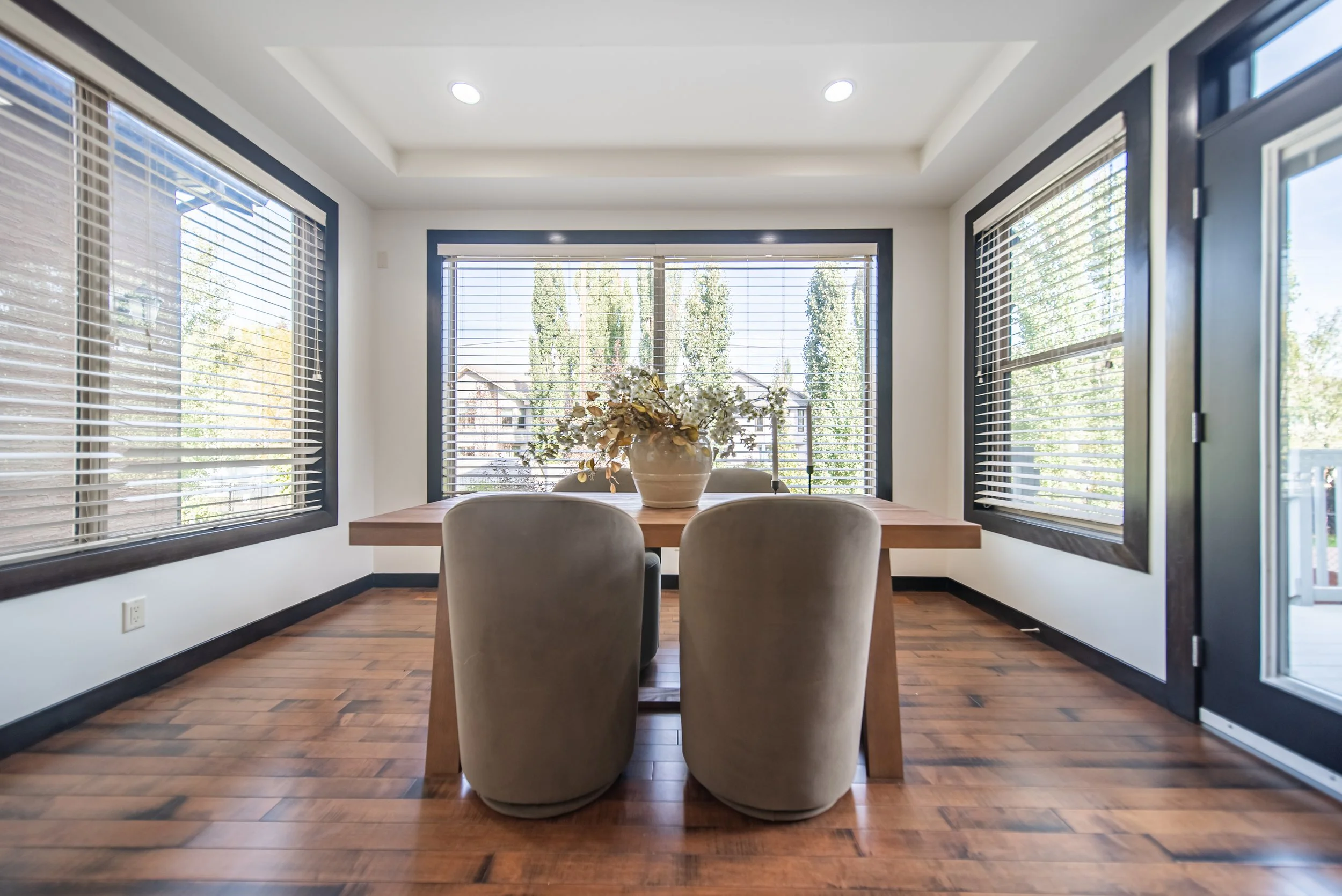 Dining room with a wooden table, two beige chairs, a vase with flowers, large windows with blinds, wooden flooring, and a door leading outside.