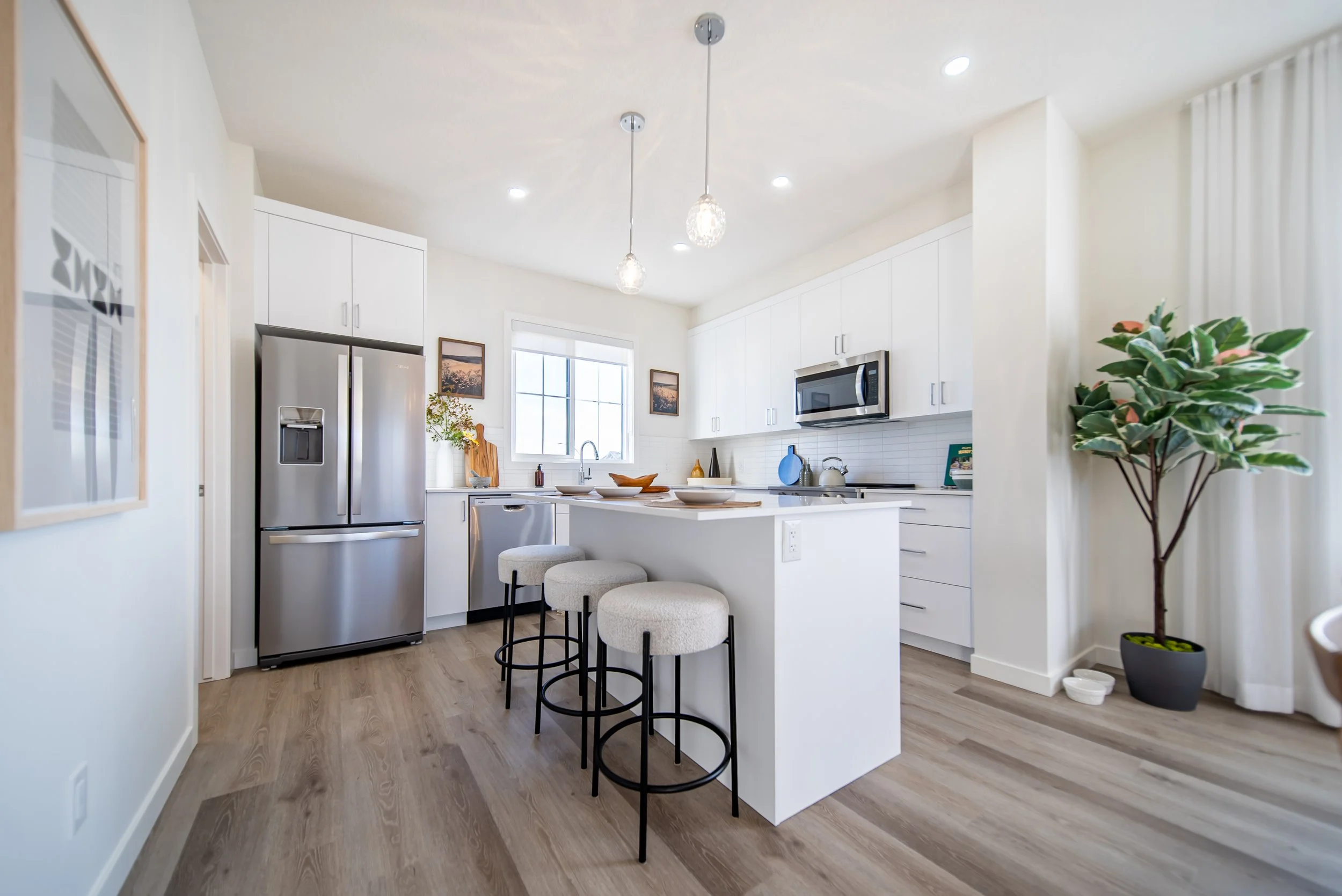 Modern white kitchen with island, gray refrigerator, microwave, and barstools, featuring wooden flooring and a large green potted plant.
