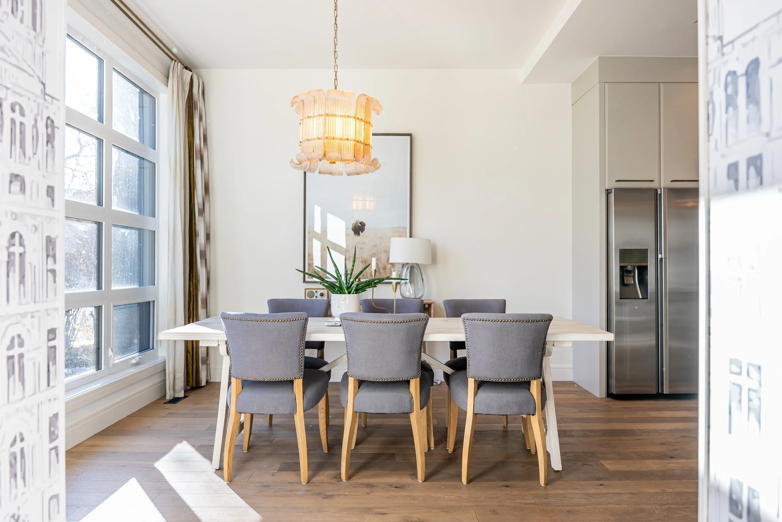 Modern dining room with six gray upholstered chairs around a white wooden table. Large windows with sheer curtains allow sunlight, and a chandelier hangs above. A plant, lamp, and framed picture are on the wall behind the table. A stainless steel ref