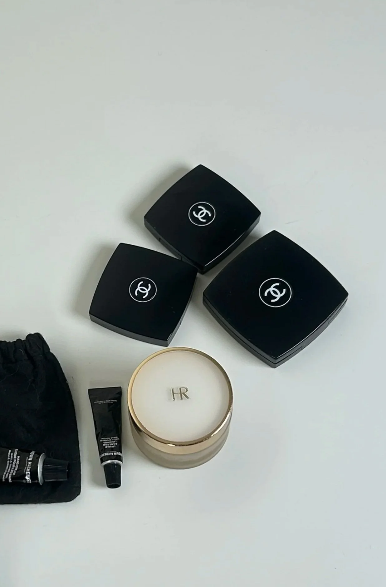 Three black Chanel powder compacts, a small black tube, a small black bottle, a cream-colored jar with gold rim and the initials HR, and a black cloth bag with a tube of skincare inside placed on a white surface.