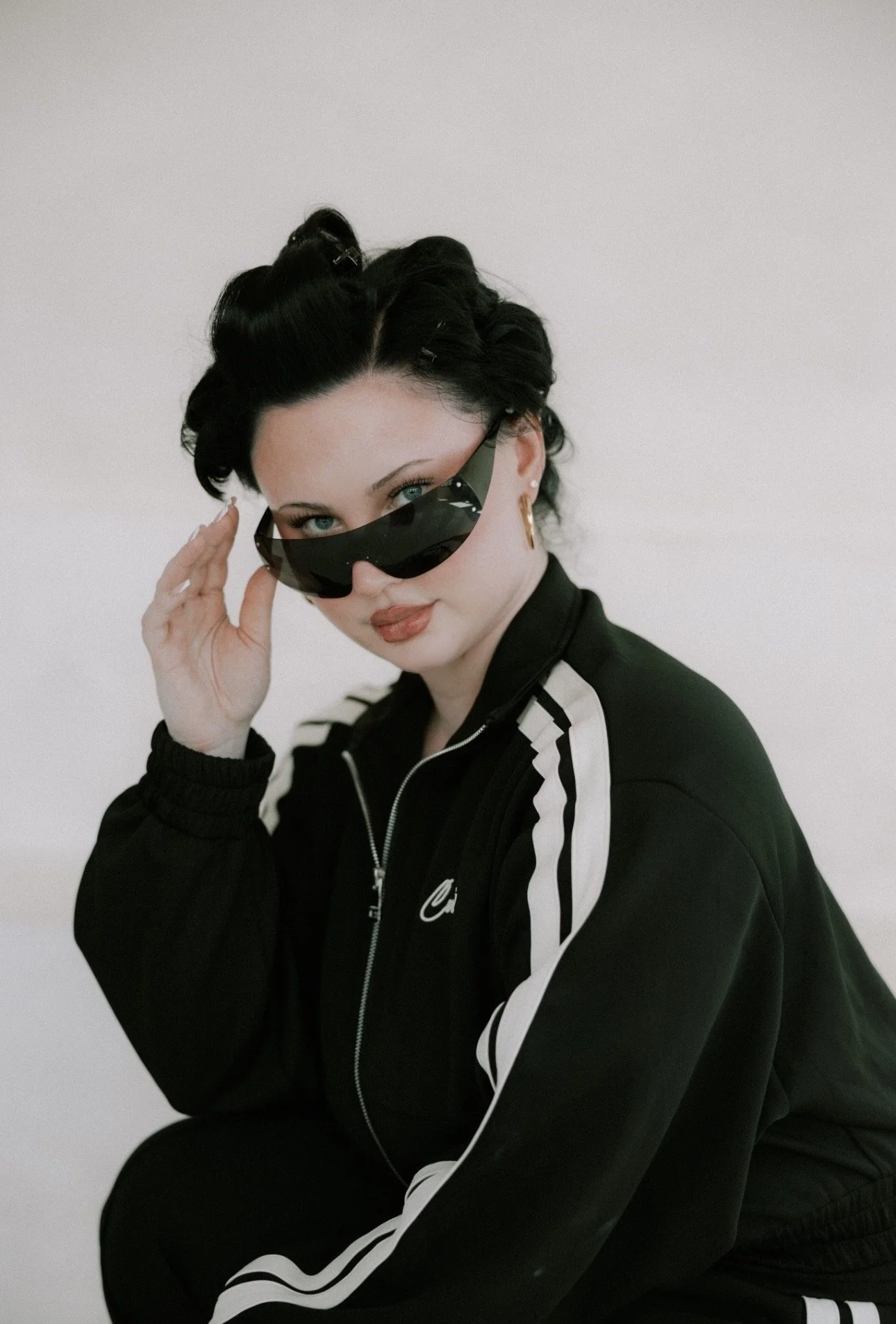 A woman with short black hair, wearing oversized sunglasses, a black Nike tracksuit with white stripes, and gold hoop earrings, posing against a neutral background.