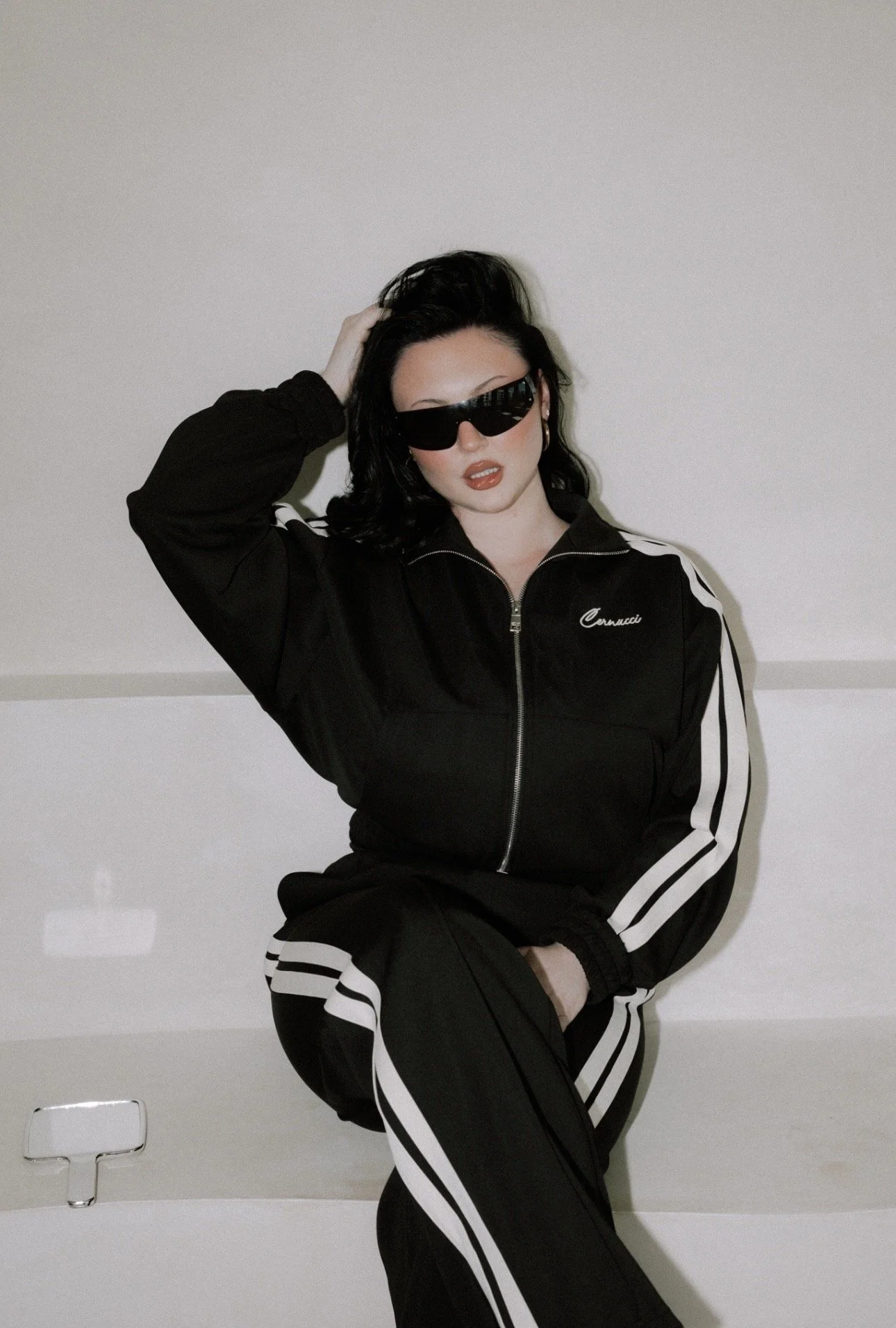 A woman with black hair and sunglasses wearing a black and white sports jacket and pants, sitting on the edge of a bathtub with a plain white wall in the background.