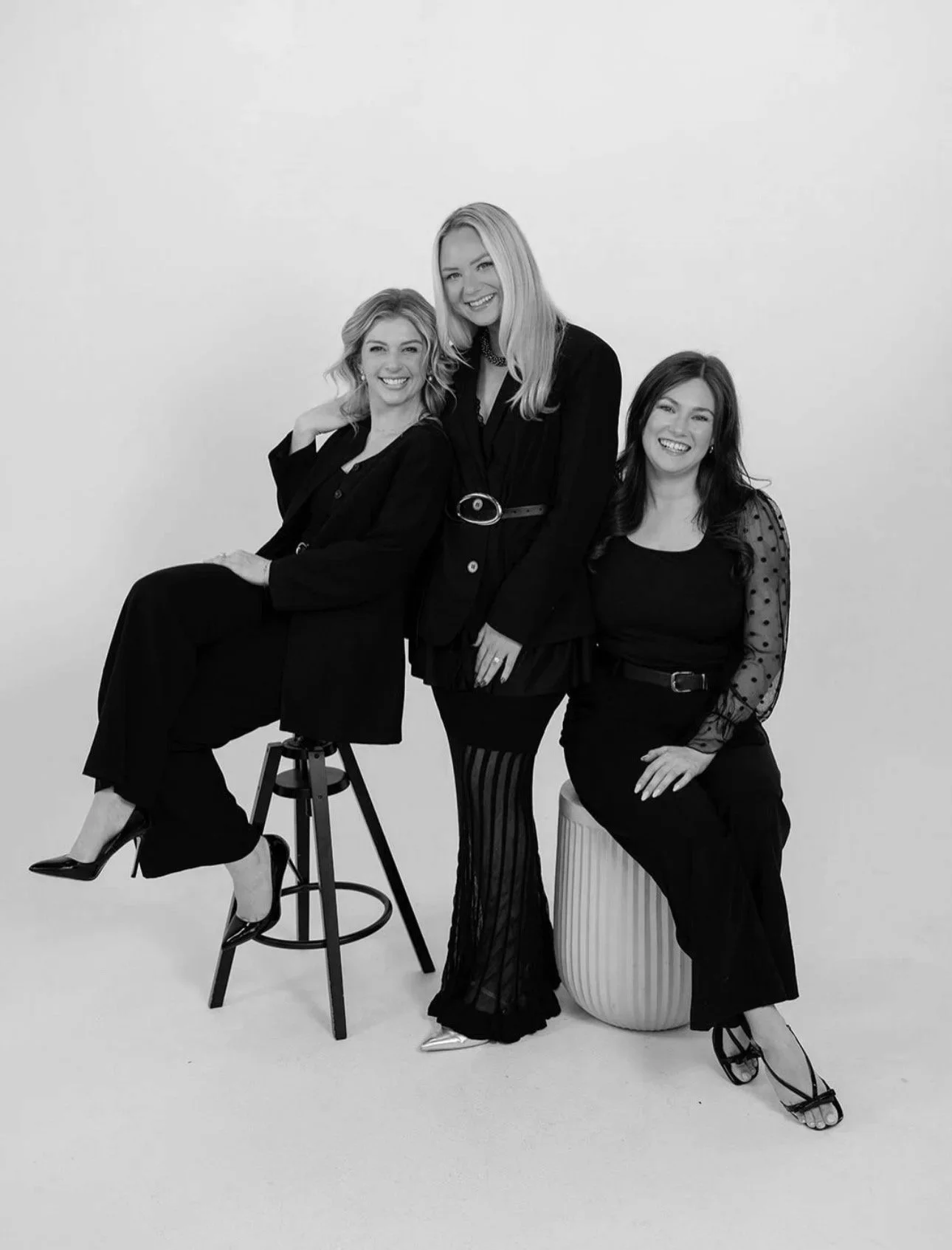 Three women in black outfits smiling, one sitting on a stool, one standing and leaning, one seated on a striped cylindrical stool, against a plain background.