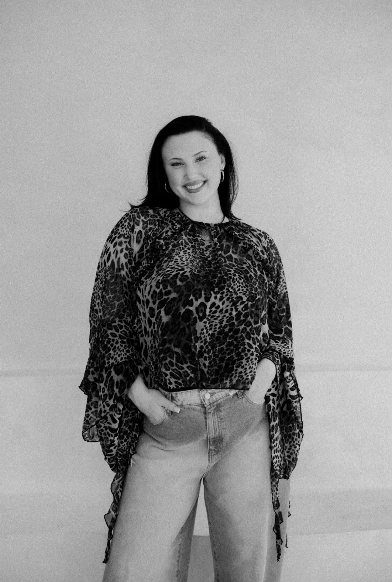A woman with dark hair and earrings smiling, wearing a leopard print blouse and high-waisted jeans, standing against a plain background.