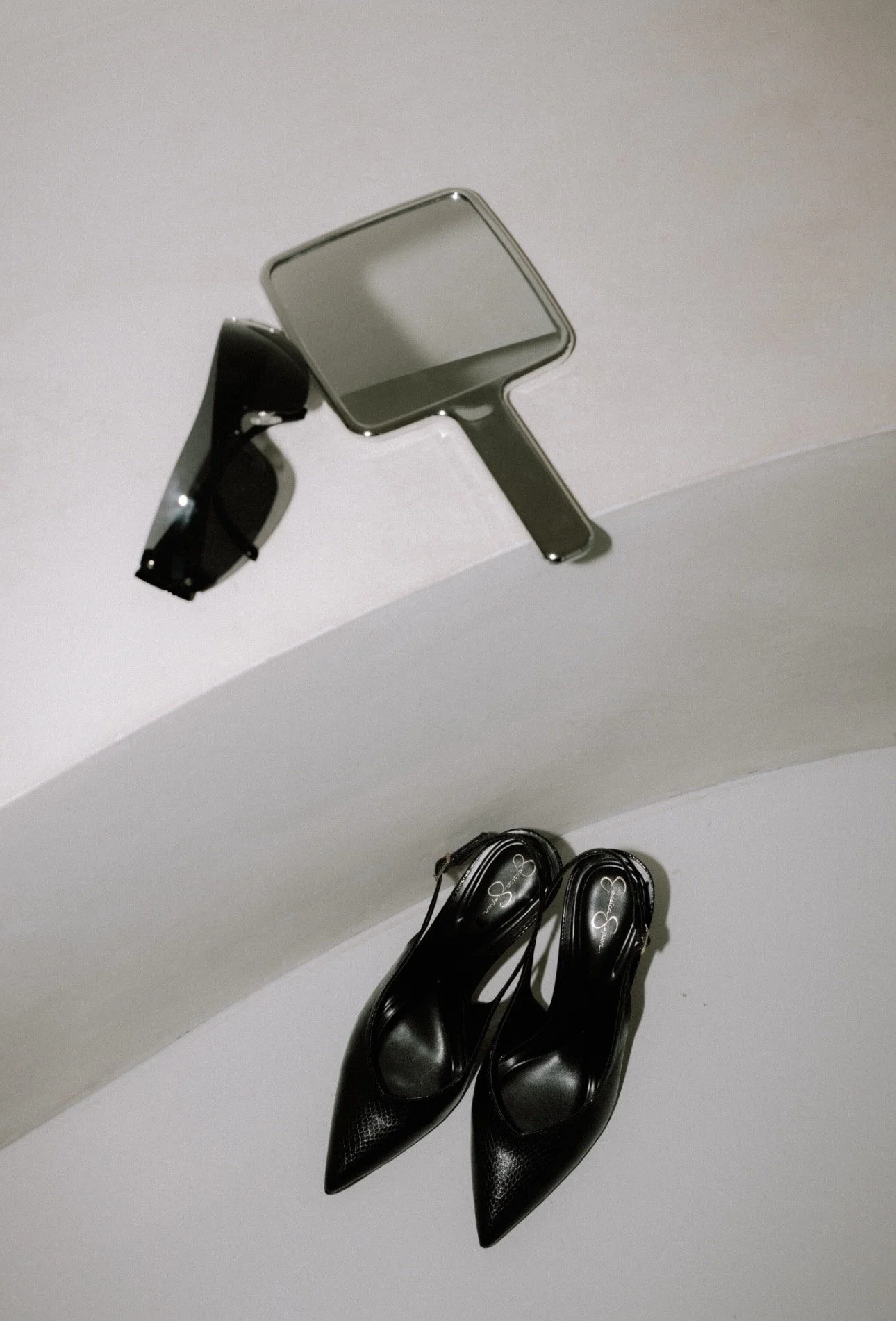 A pair of black high-heeled shoes, a mirror, and sunglasses on a curved white surface.