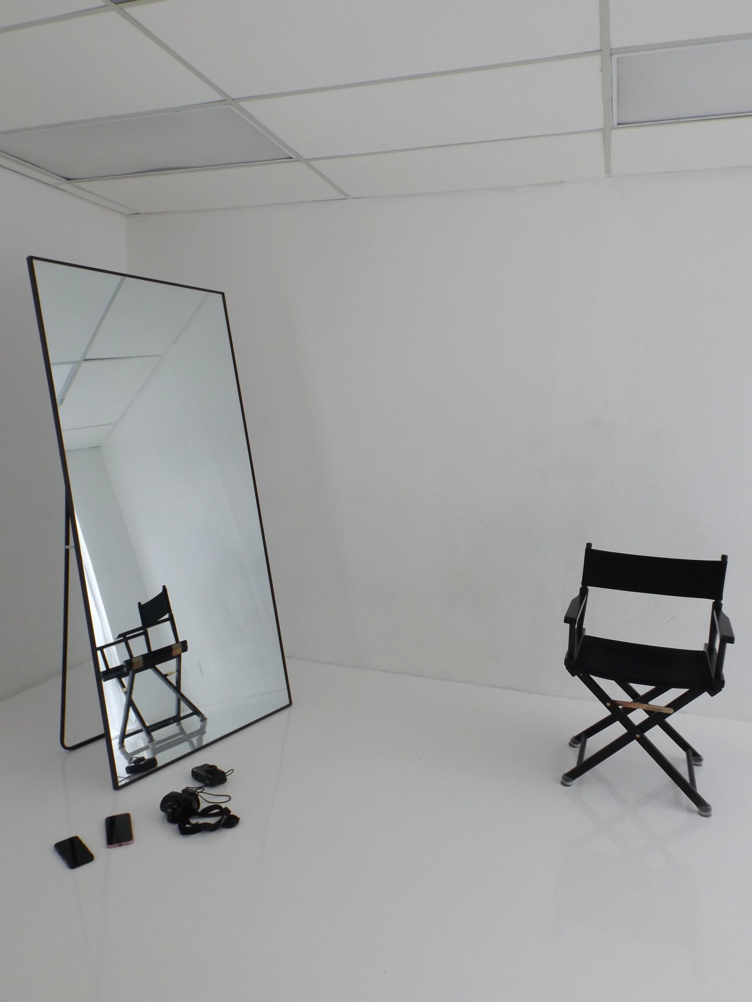 A makeup or photography studio with a black director's chair, a large tilted mirror, and some small objects on the floor including two phones, a camera, and a strap, all in a white room with ceiling panels.