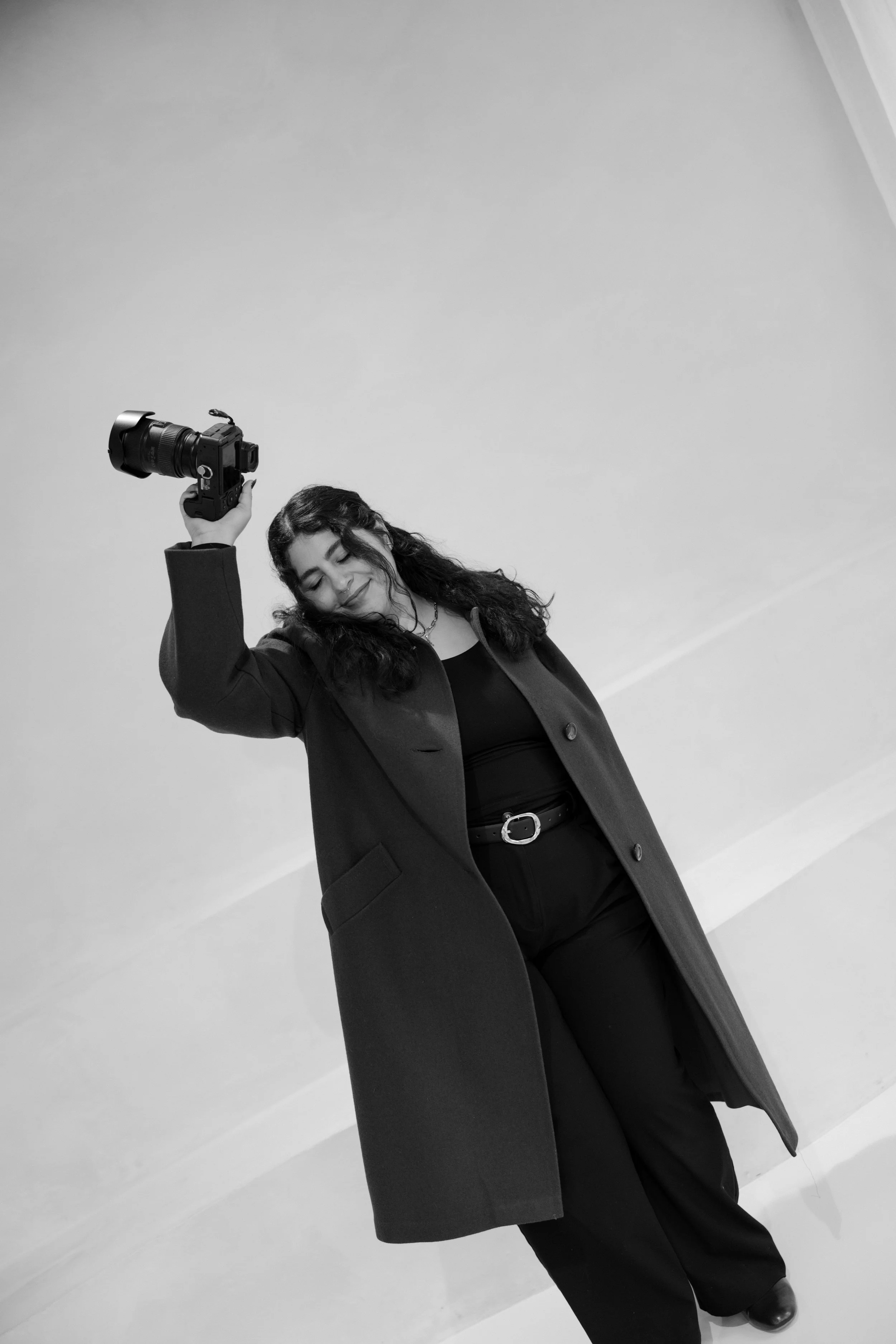 A woman with curly hair smiling and holding a camera above her head, standing against a plain wall.