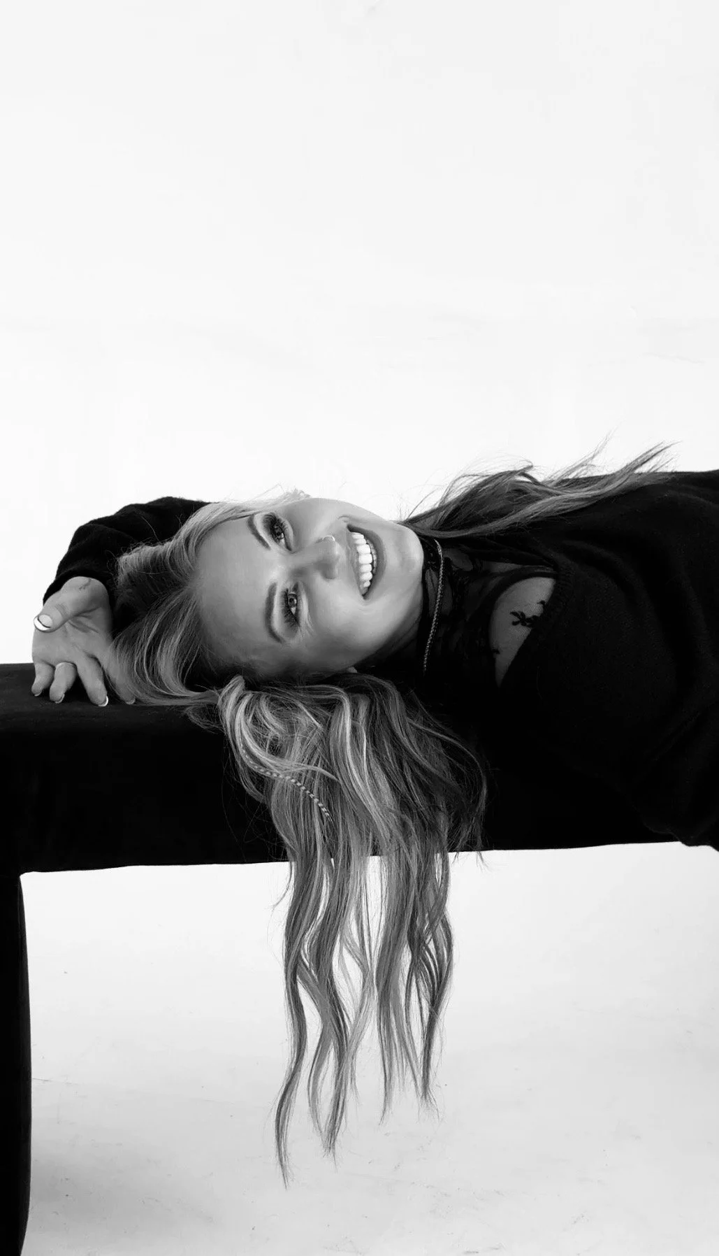 Black and white photo of a woman with long wavy hair, smiling and leaning on a table with her head resting on her arm.
