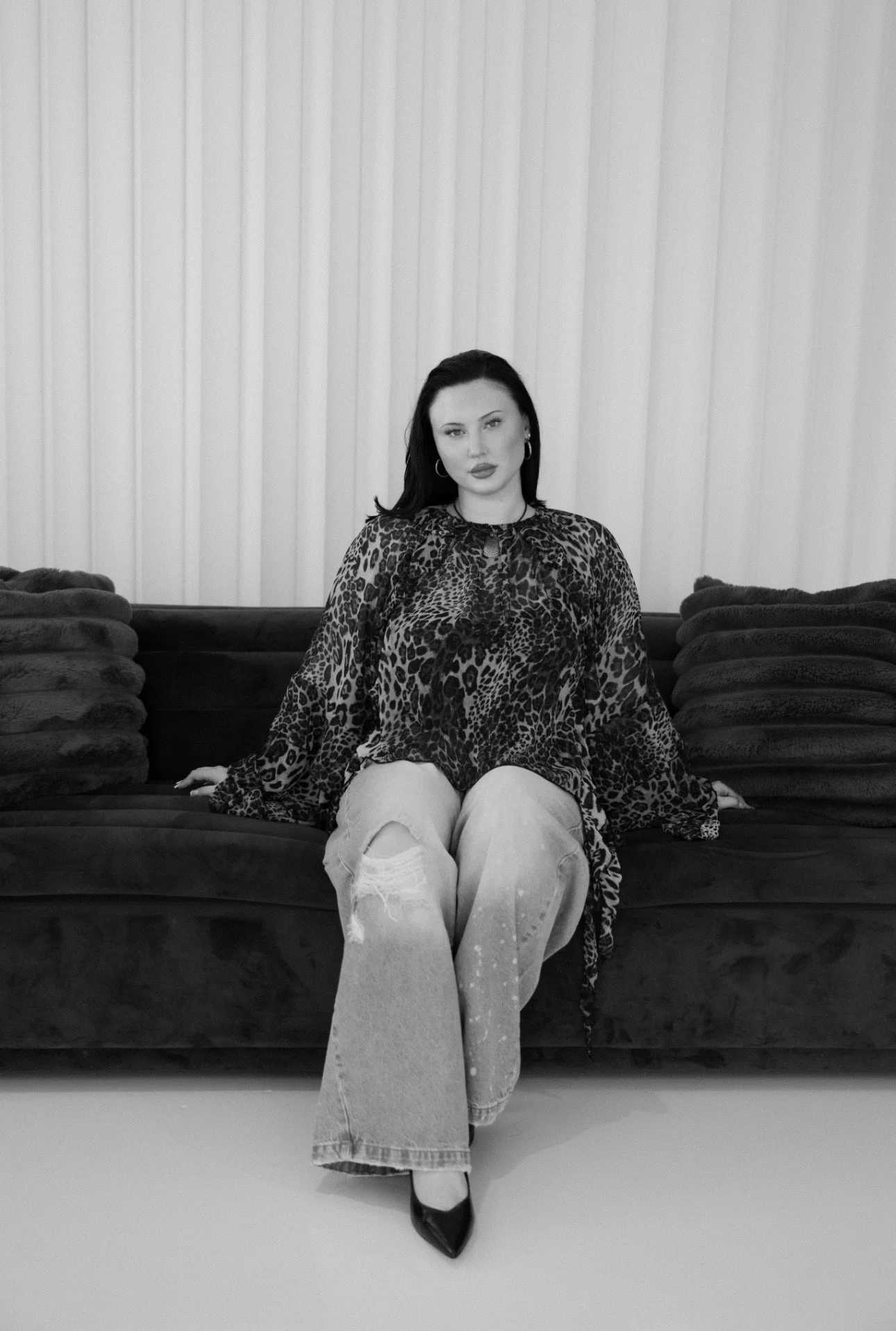 A woman sitting on a dark sofa with multiple pillows, wearing a leopard print top, ripped jeans, and black heels, in front of vertically striped curtains.