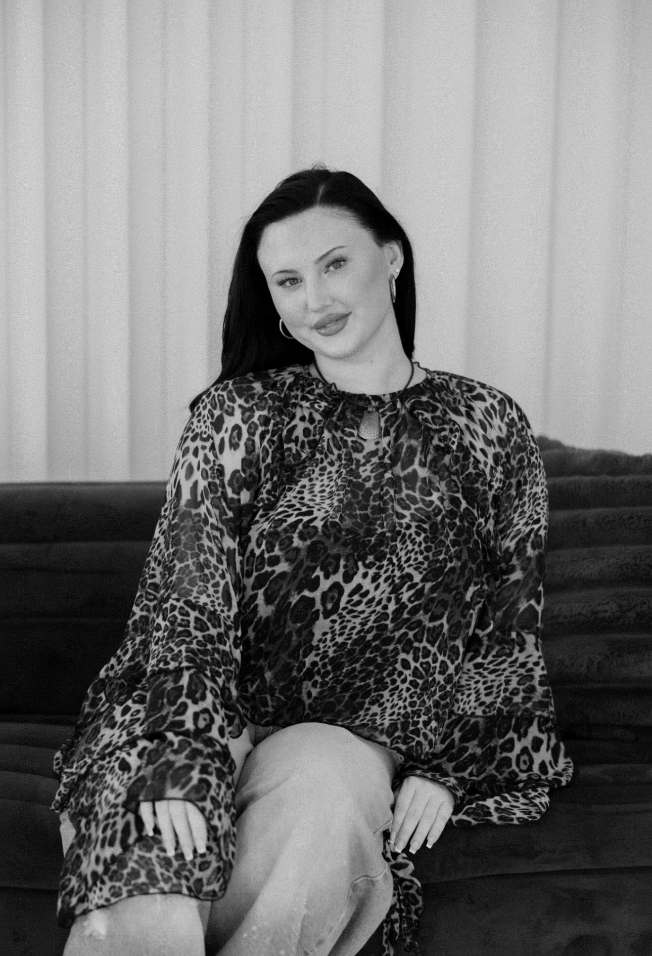 A woman with black hair and hoop earrings sitting on a couch, wearing a leopard-print blouse and light-colored pants.