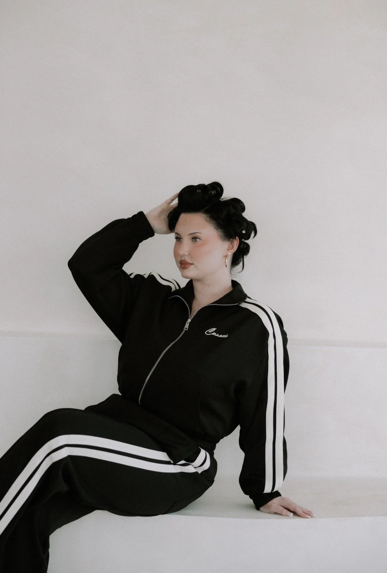 A woman with vintage hairstyle wearing a black tracksuit with white stripes, sitting against a plain white background.