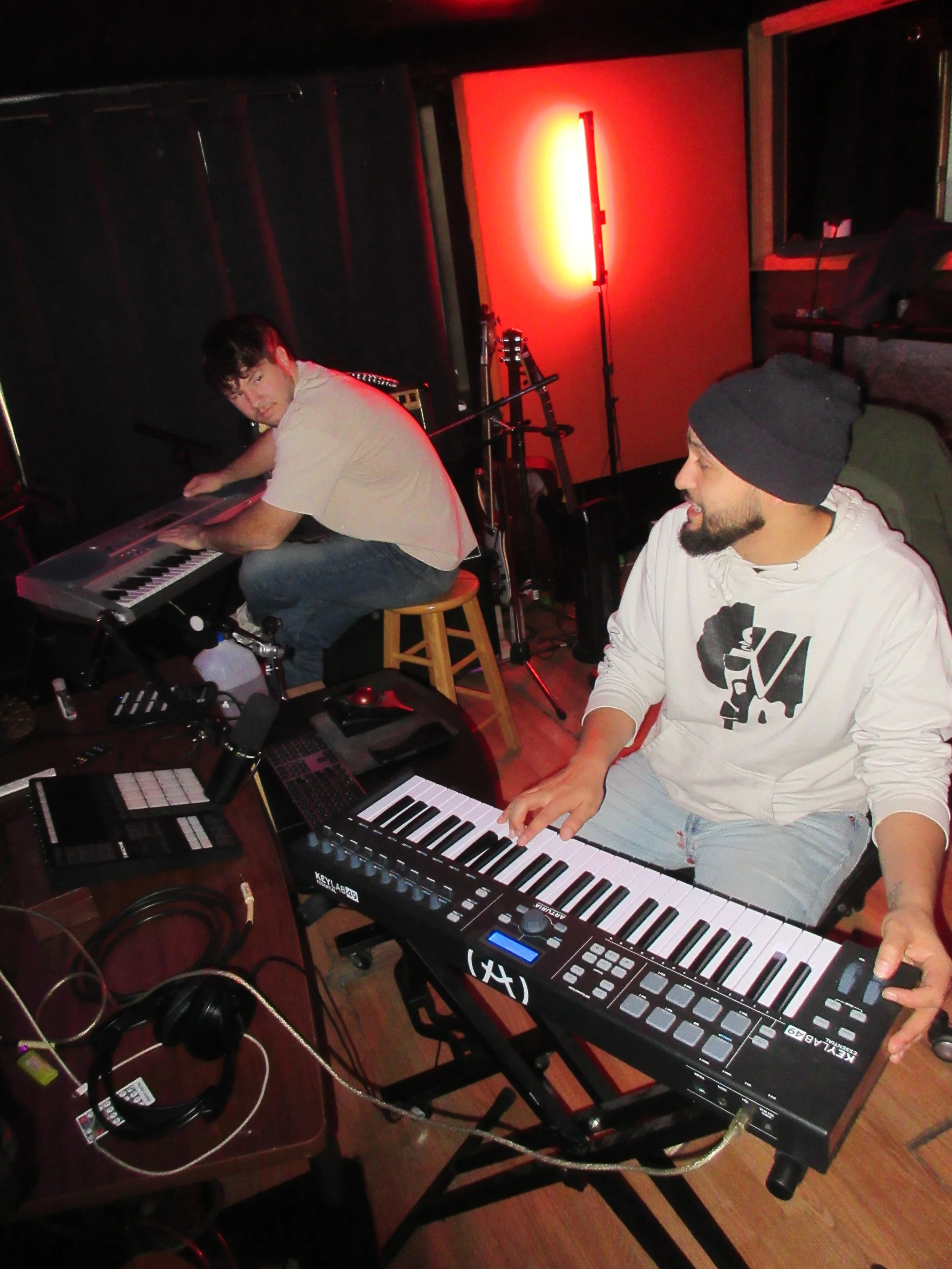 Two musicians in a recording studio, one playing a keyboard and the other sitting on a stool, with musical equipment and red lighting in the background.