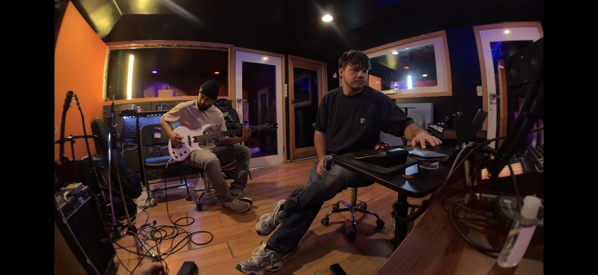 Two men in a music studio, one playing an electric guitar and the other working with audio equipment at a desk.