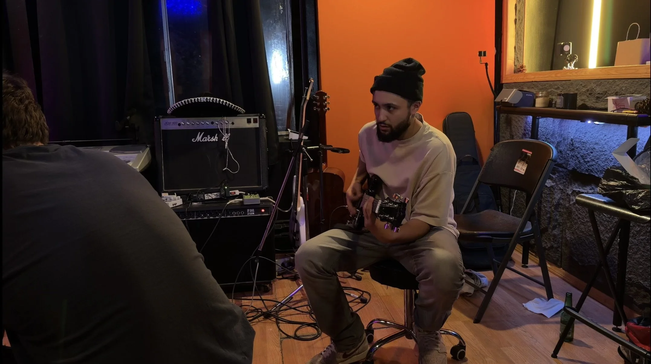 A man with a beard wearing a black beanie and beige t-shirt playing an electric guitar on a stool in a music studio with orange and textured walls, surrounded by musical equipment and chairs.